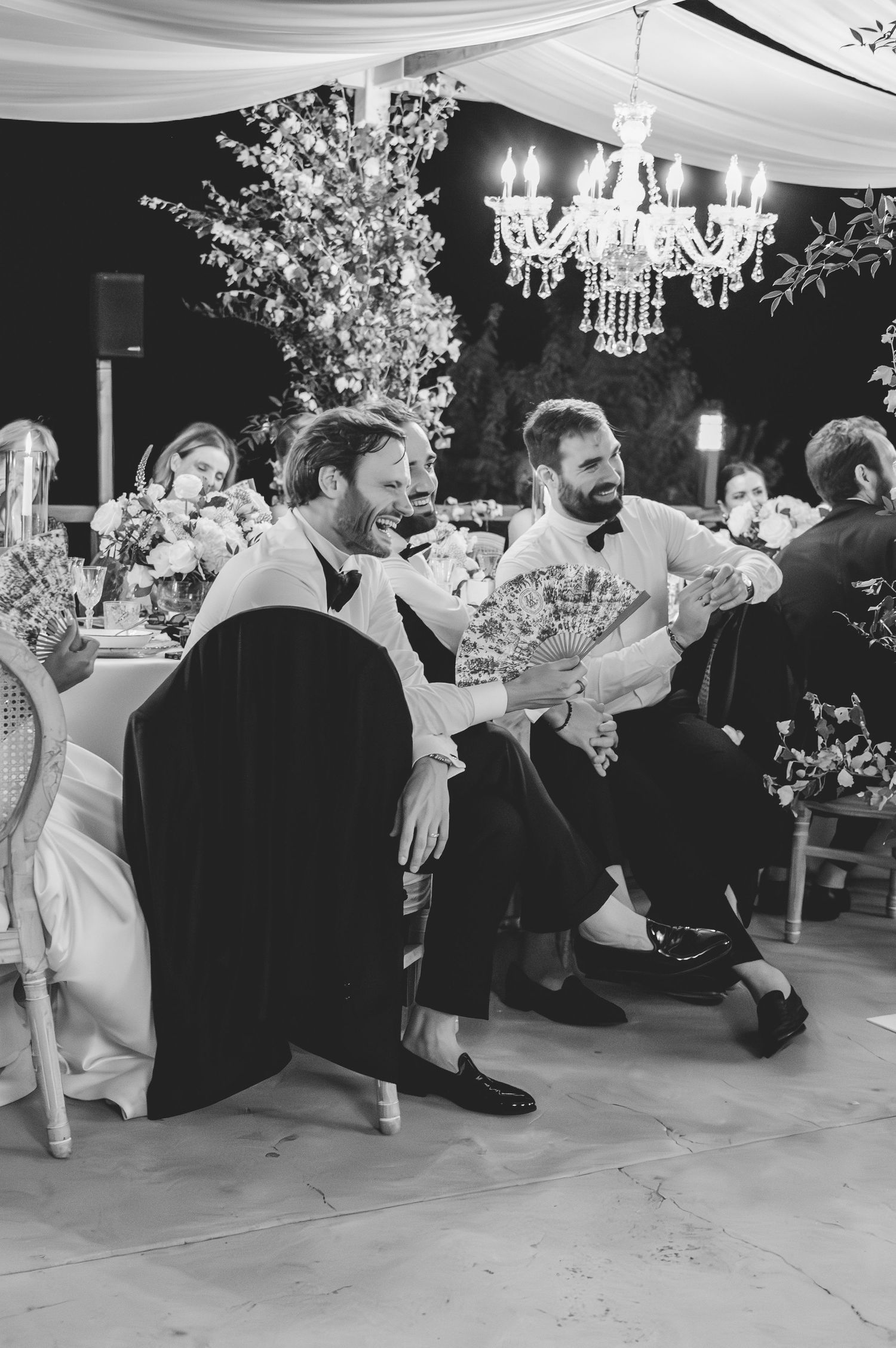 Black and white photo of two groomsmen laughing and holding decorative fans at elegant wedding reception.