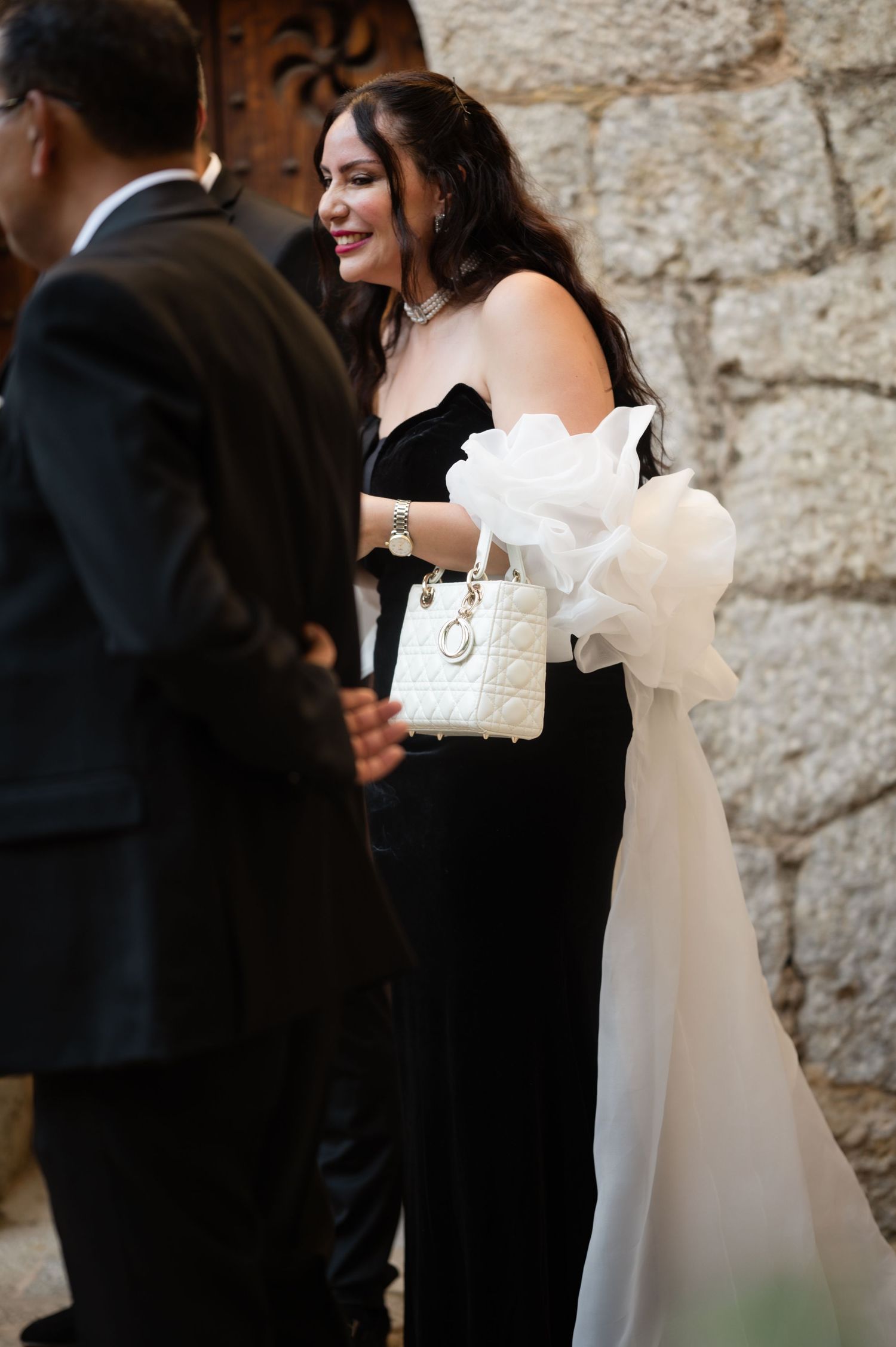 Woman in black strapless gown with white organza bow carries a white Lady Dior bag at a wedding.