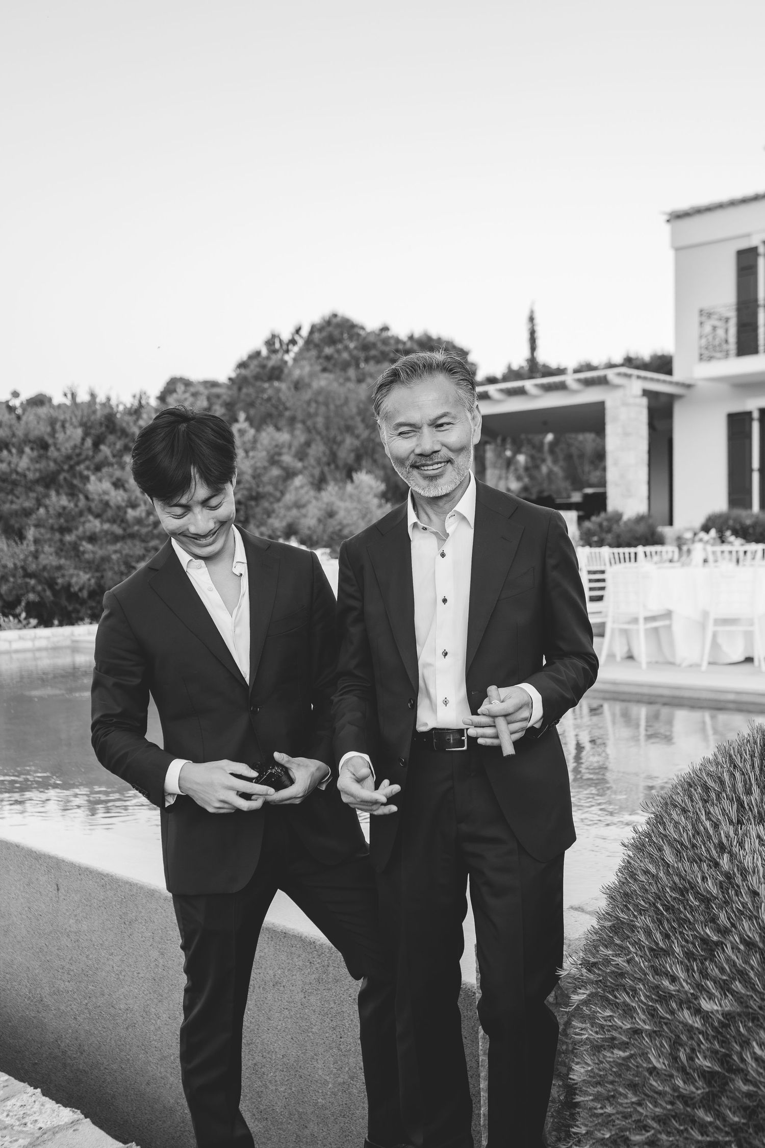 Two men in dark suits smile and chat outdoors near a pool at a wedding venue in black and white.