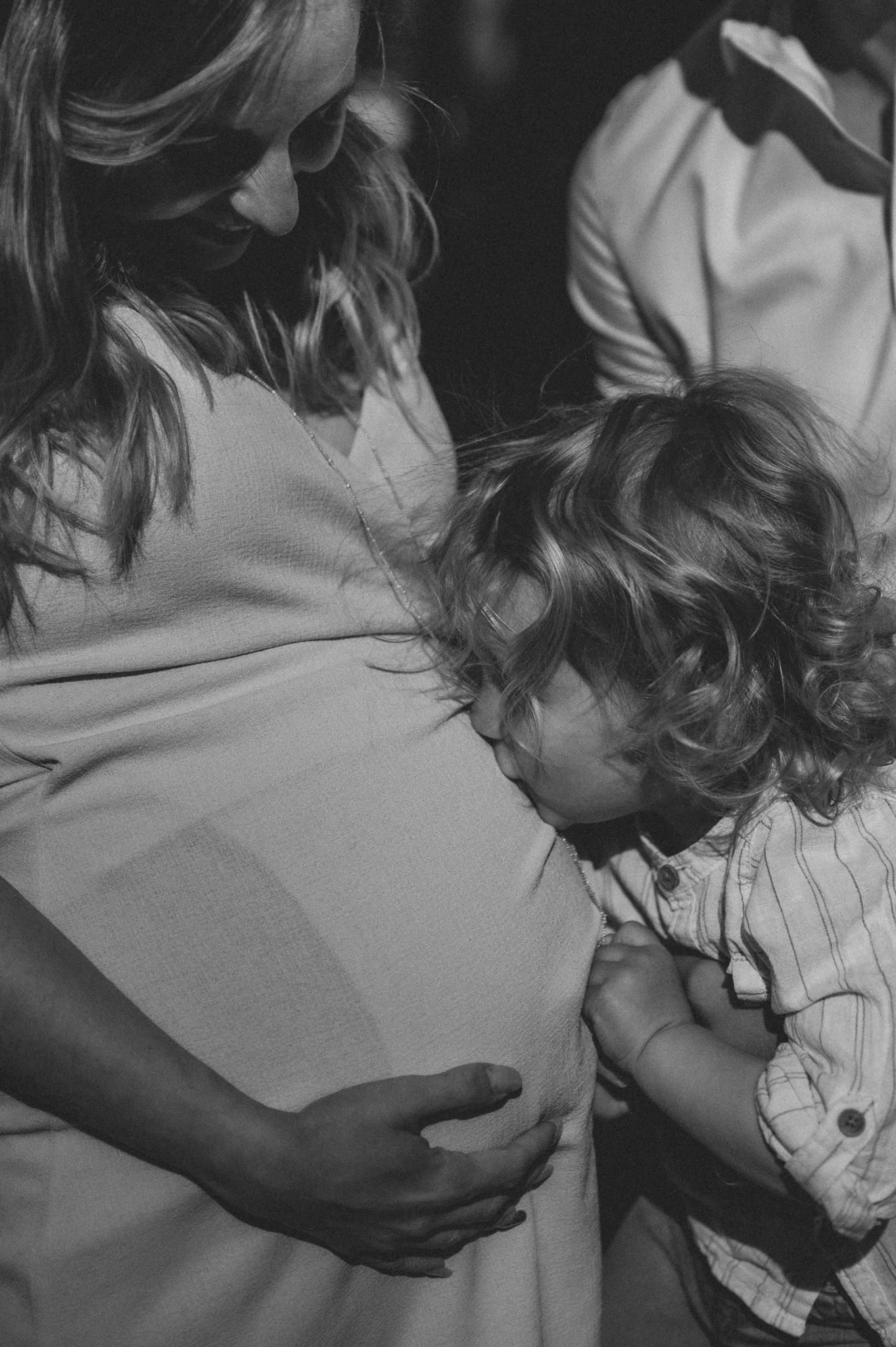 Black and white photo of a toddler kissing a pregnant woman's belly, a tender family moment.