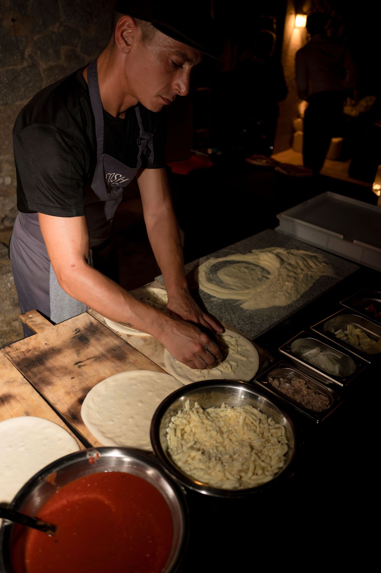 Chef preparing fresh pizza dough with toppings at a dark wedding catering station.