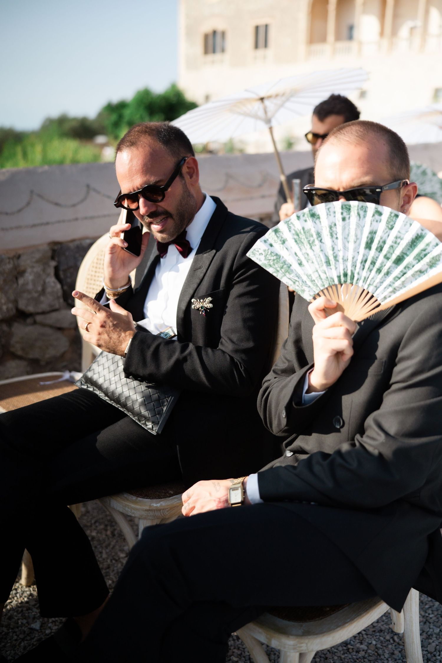 Two men in black suits sit outdoors at a wedding, one on phone, one holding a decorative fan.