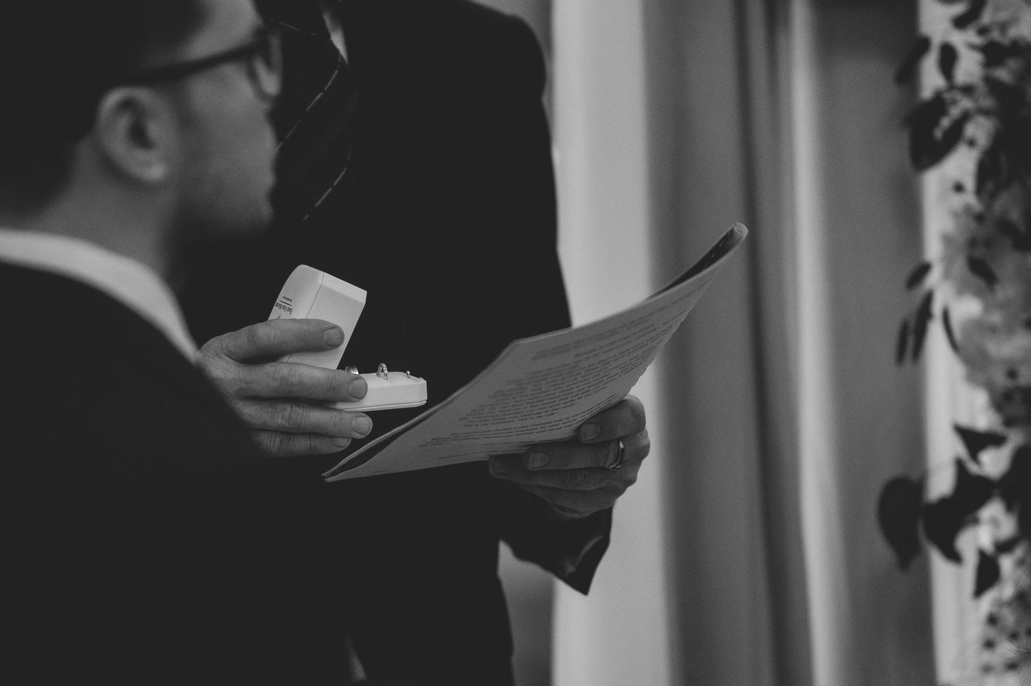 Black and white image of a man holding a ring box and reading vows from a paper.