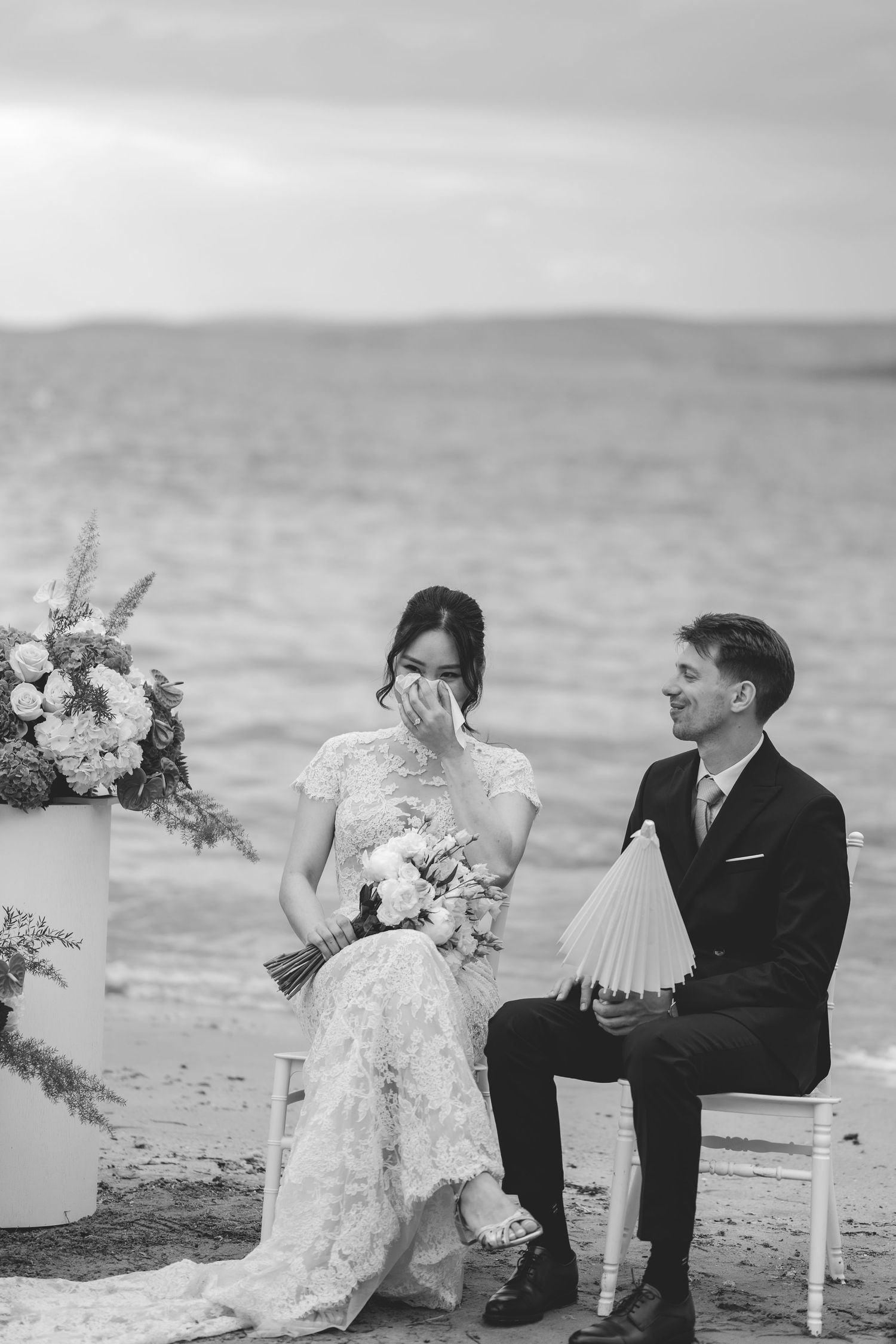 Emotional black and white photo of bride wiping tears during beach wedding ceremony.