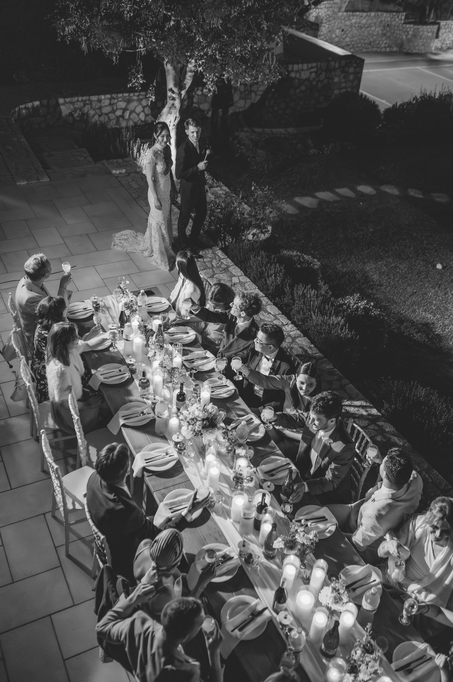 Aerial black and white photo of wedding guests gathered around a candlelit outdoor dinner table at night.