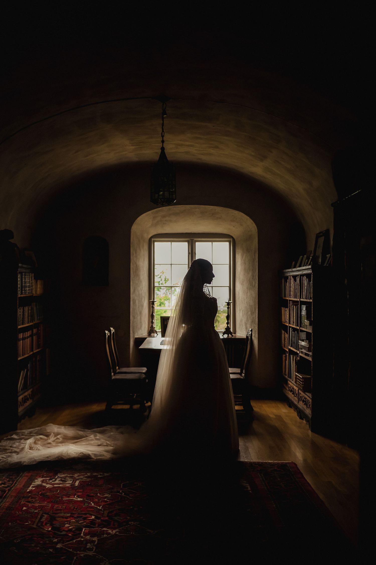 Silhouette of bride in cathedral veil standing before arched window in dark library room.