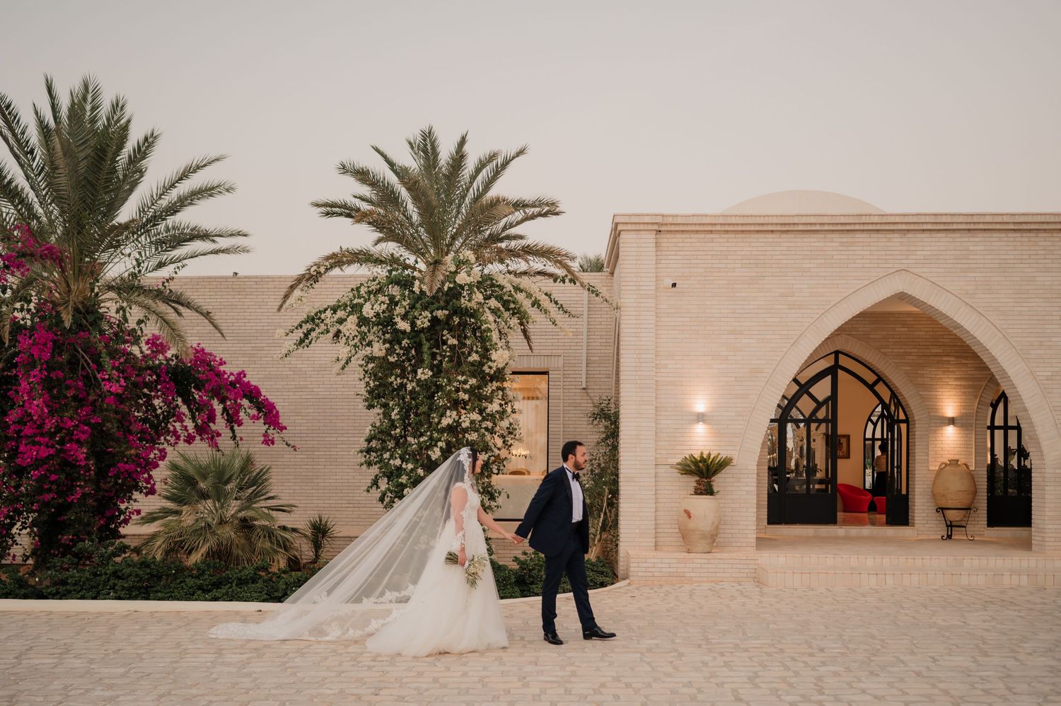 Wedding couple walking hand in hand past ornate stone archway with bougainvillea and palm trees at dusk.
