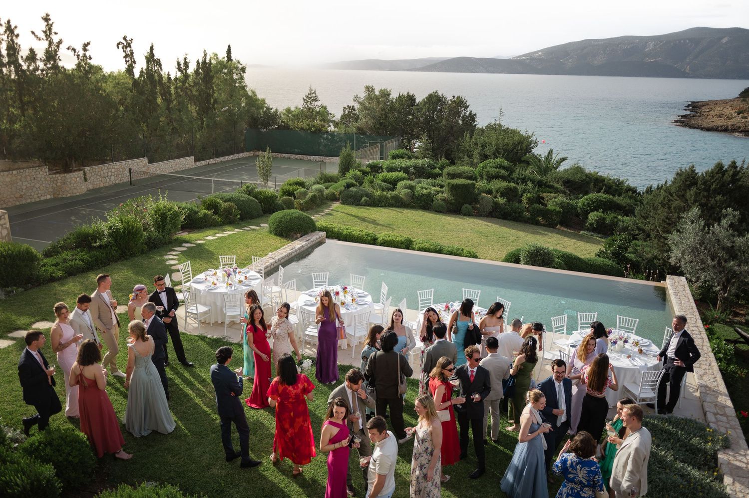 Wedding guests mingle at elegant outdoor reception beside infinity pool with sea views.