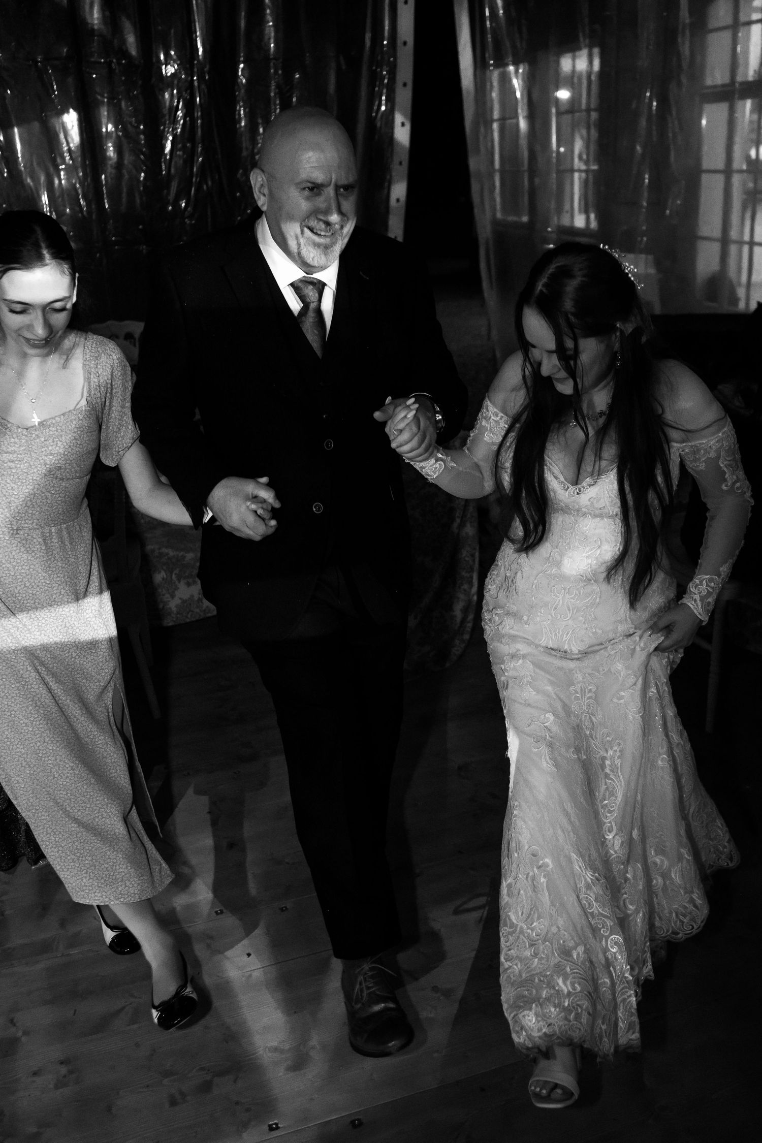 Bride and guests laugh while dancing together at wedding reception in black and white photo.