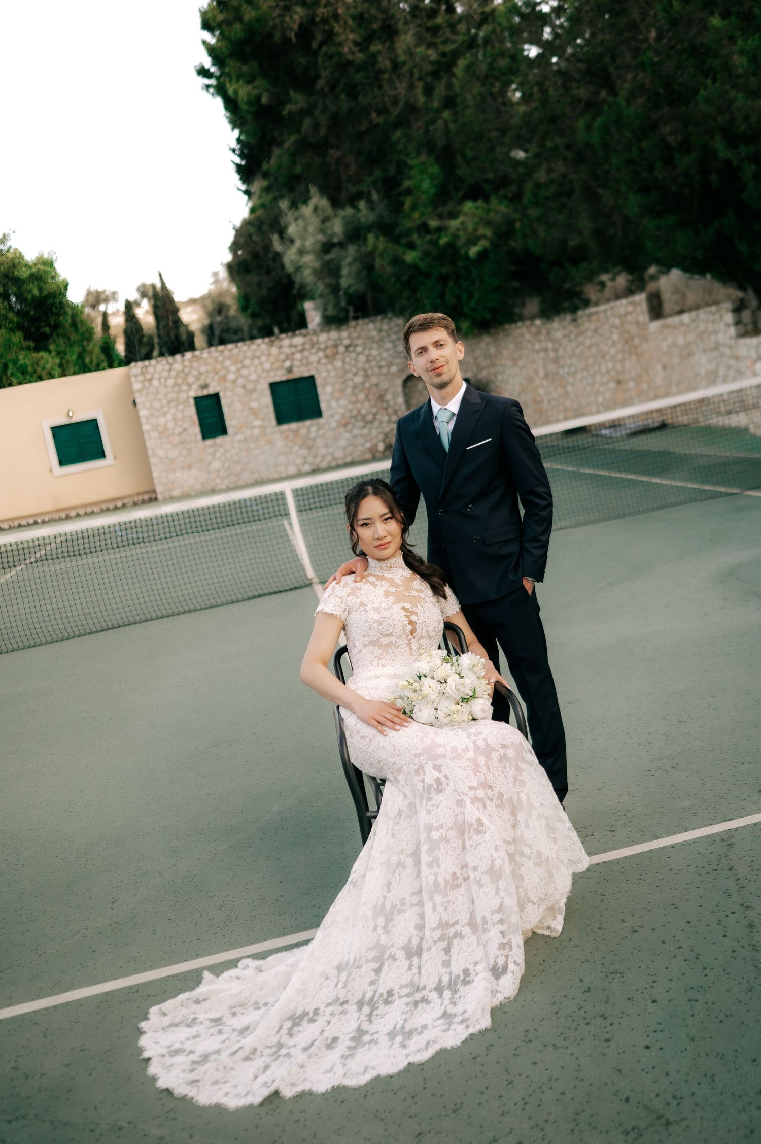 Bride in lace gown and groom in navy suit pose on tennis court at wedding.