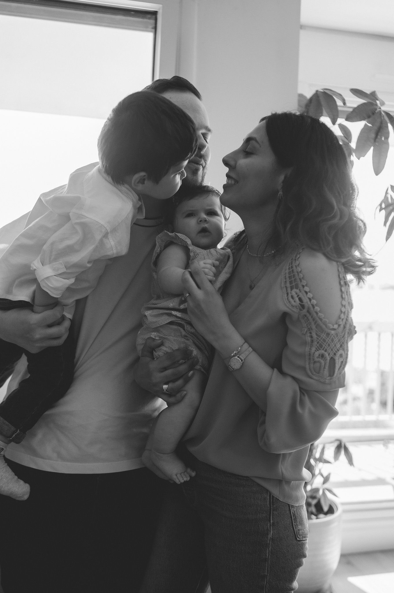 Black and white family portrait of parents holding two young children near a bright window indoors.