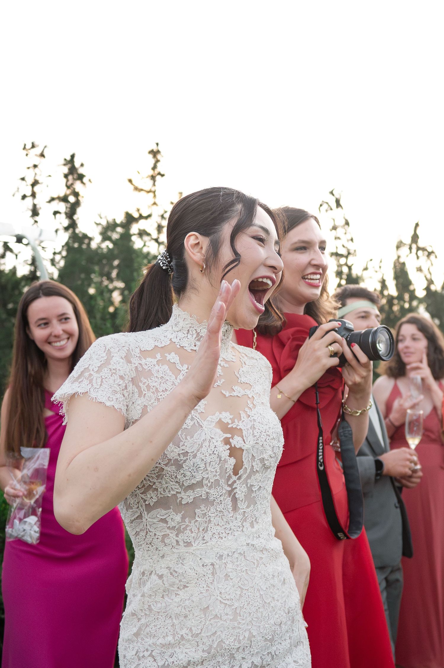 Laughing bride in white lace dress reacts with joy alongside guests and photographer at outdoor wedding.