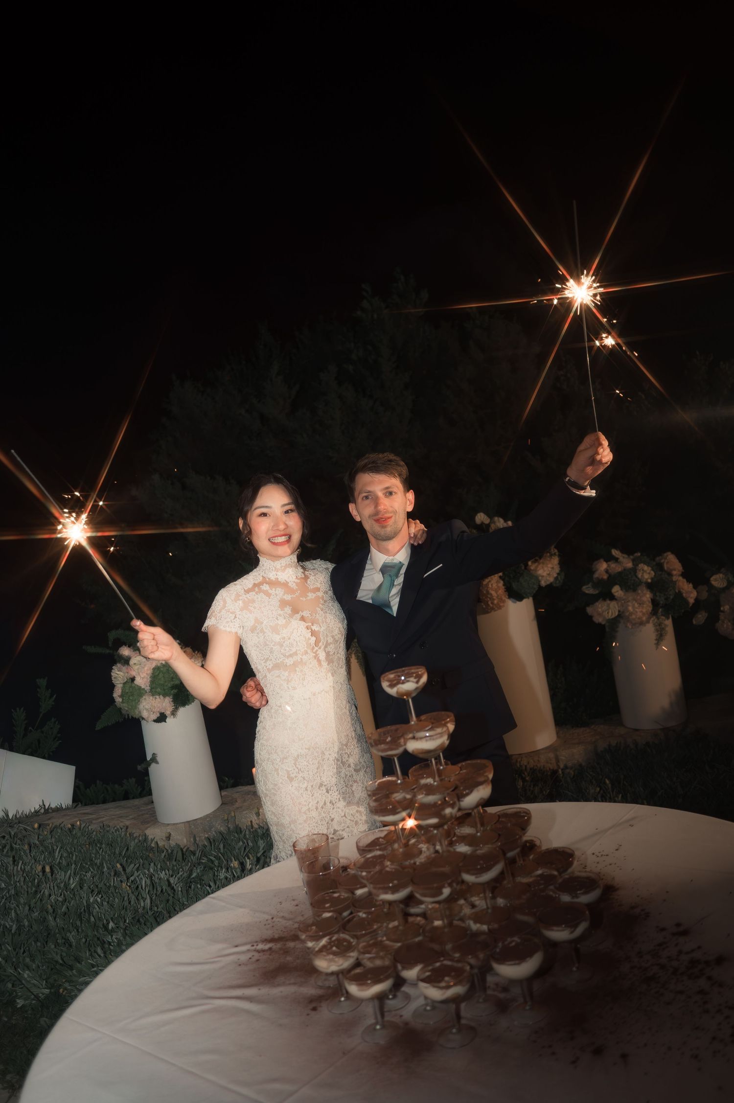 Smiling bride and groom holding sparklers beside a champagne tower at their outdoor wedding.