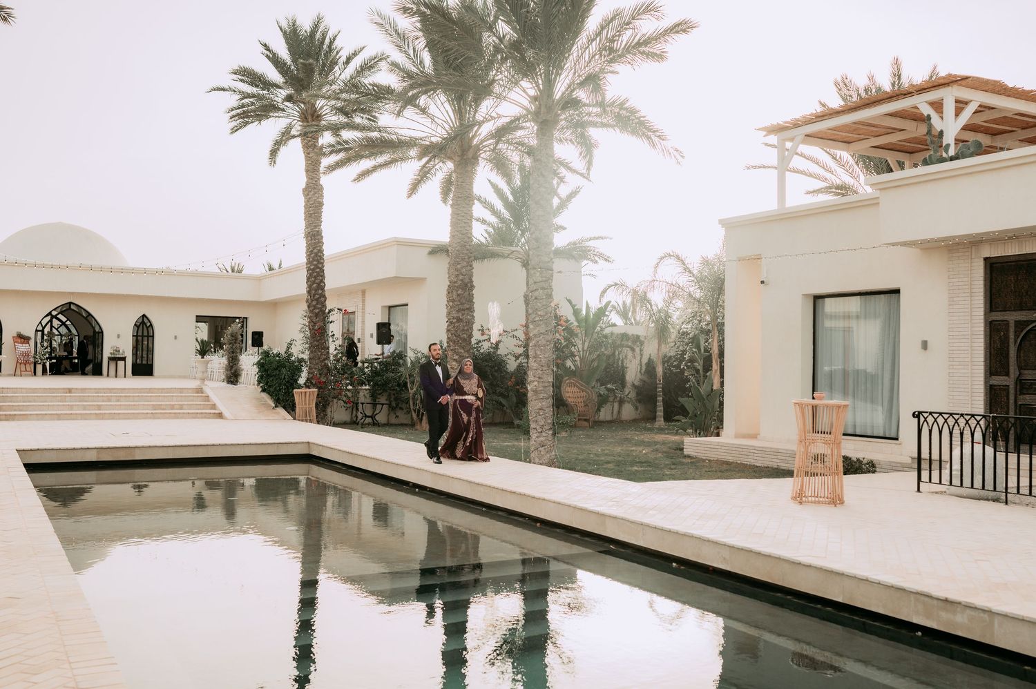 Couple strolling beside a tranquil reflecting pool at a white stucco wedding venue surrounded by palm trees.