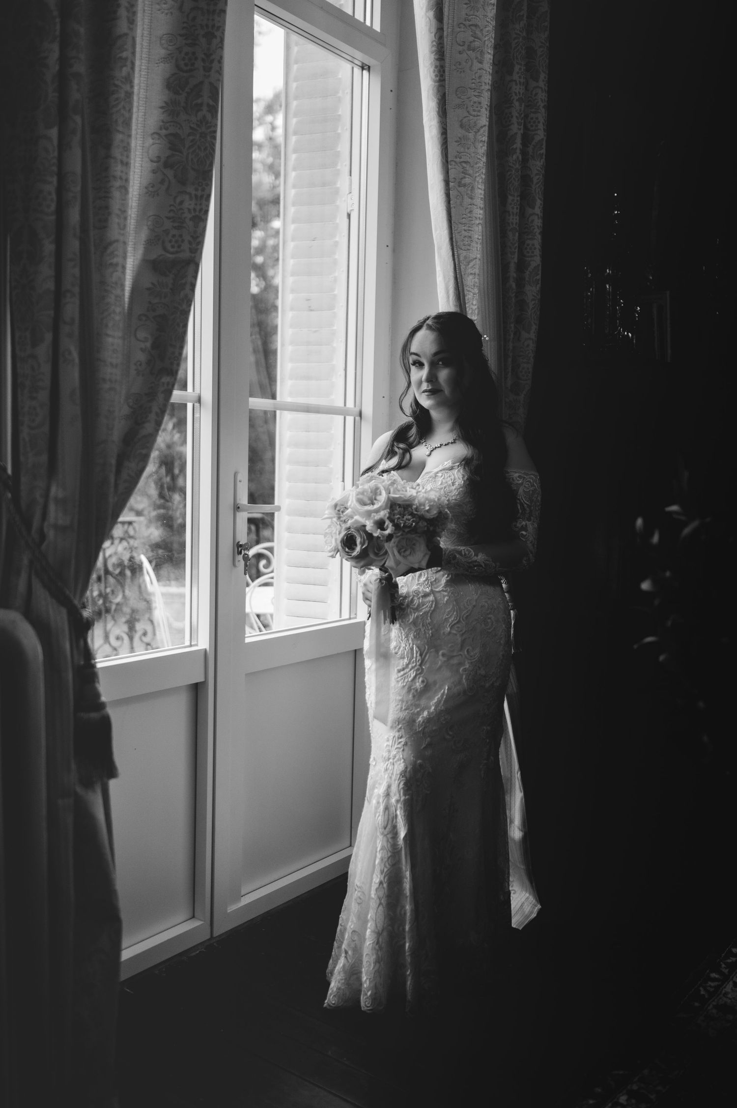 Black and white portrait of a bride in lace gown holding bouquet while standing by a tall window.