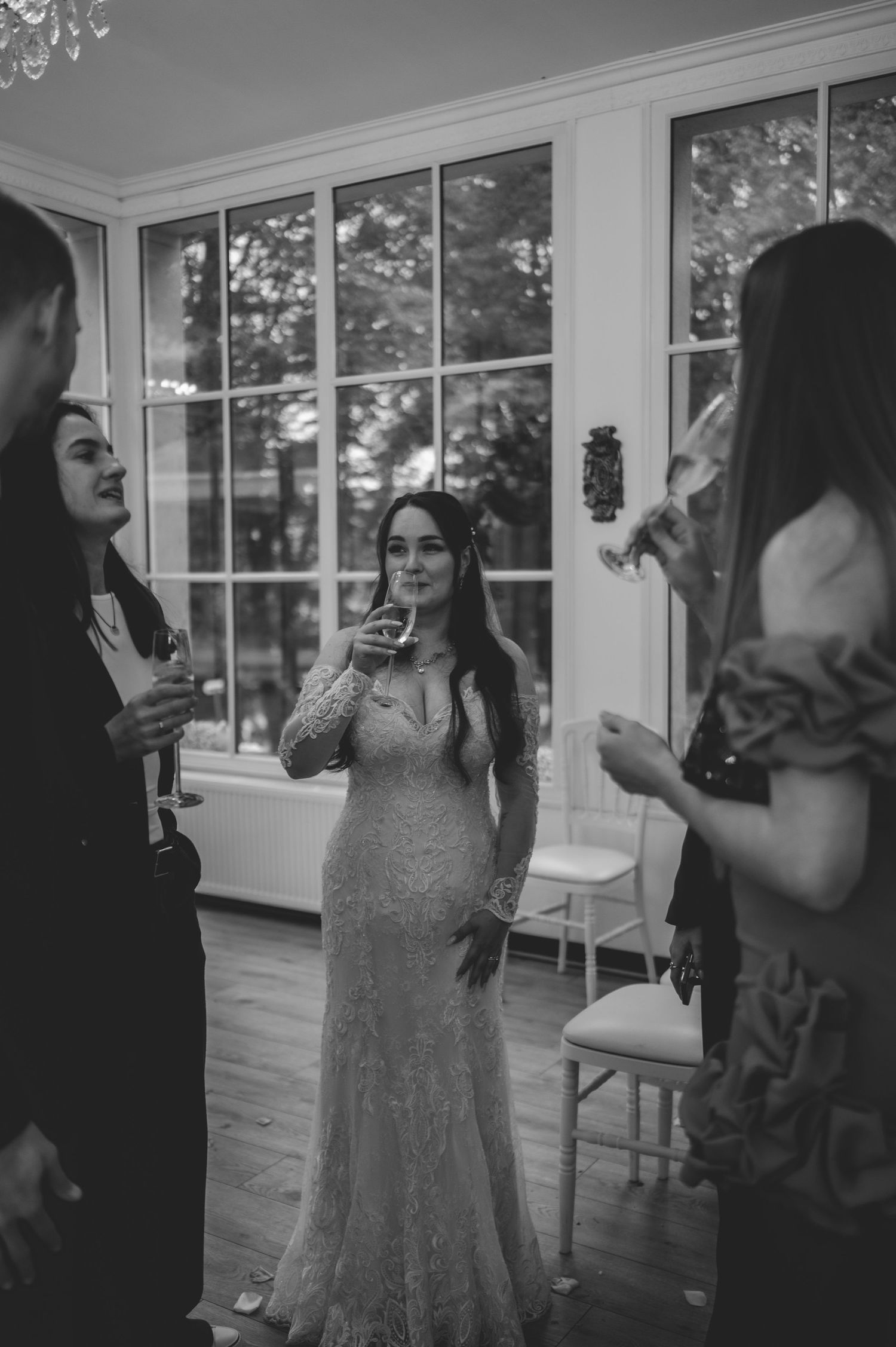 Bride in lace wedding gown holding champagne glass at indoor reception near large windows.