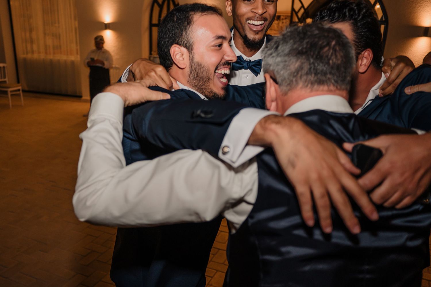 Groom shares joyful group hug with friends in suits at wedding reception celebration.
