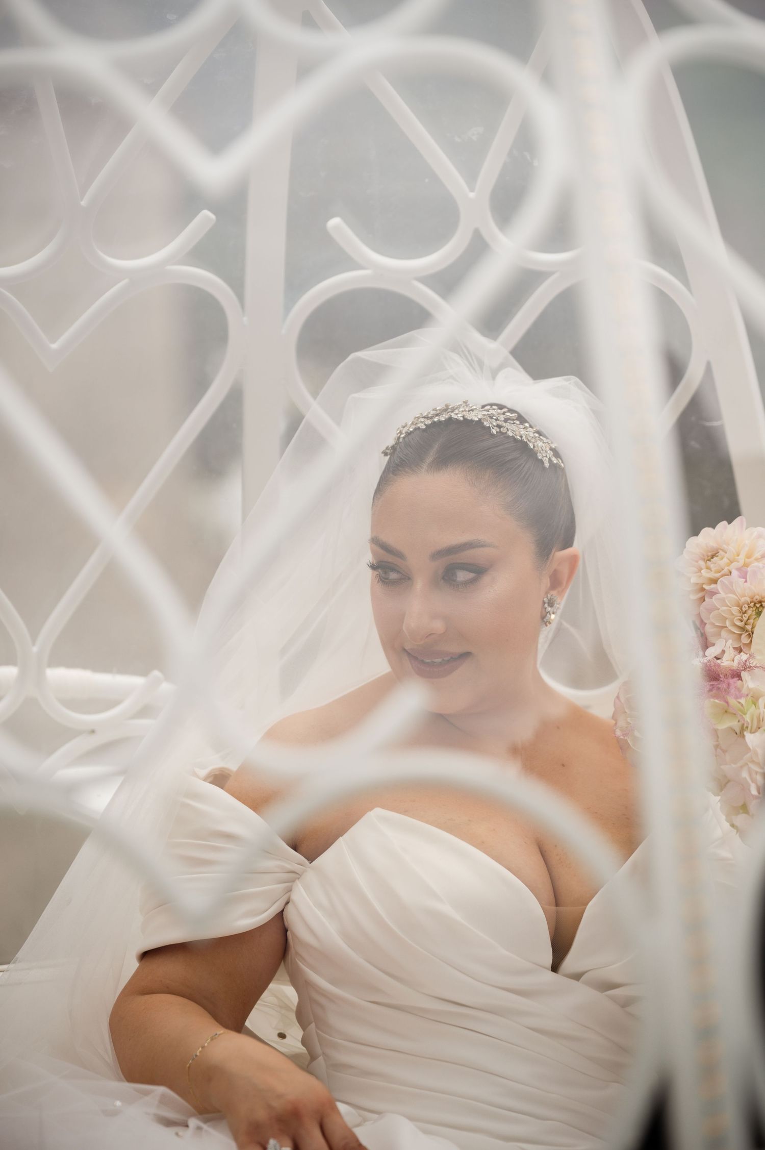 Beautiful bride wearing crystal tiara and veil holding pink bouquet, photographed through decorative white lattice.