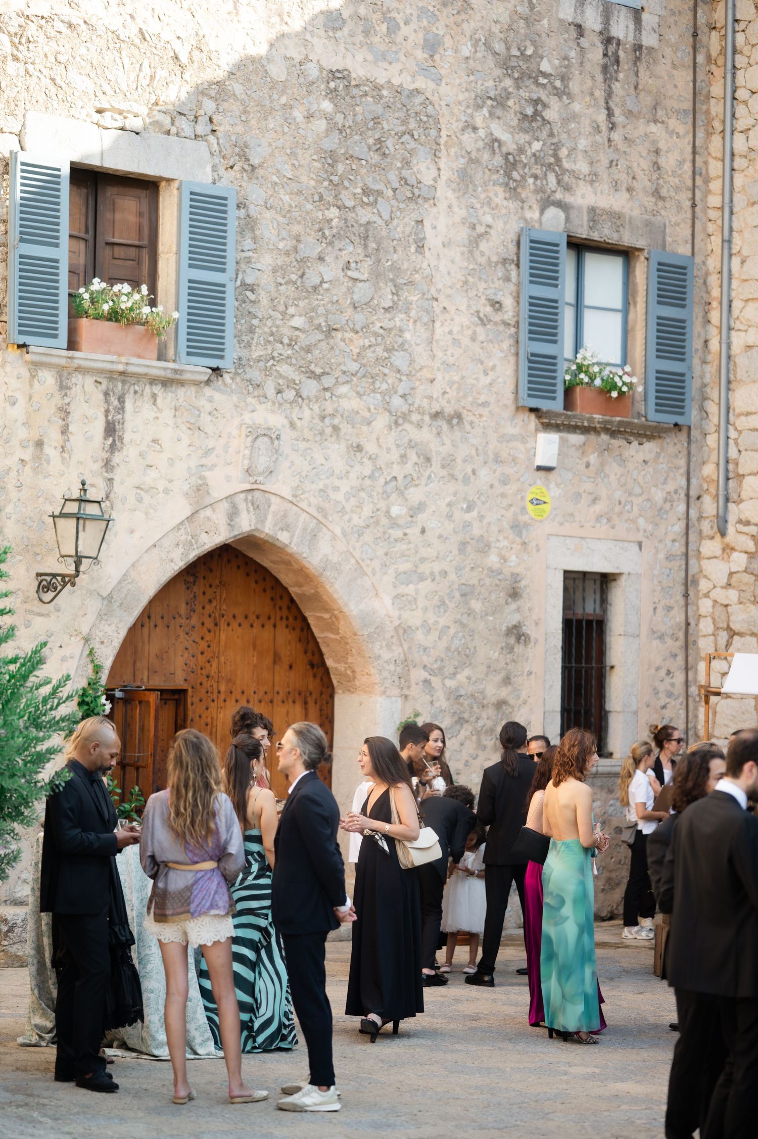 Elegantly dressed wedding guests mingle outside a rustic stone building with arched doorway and blue shutters.
