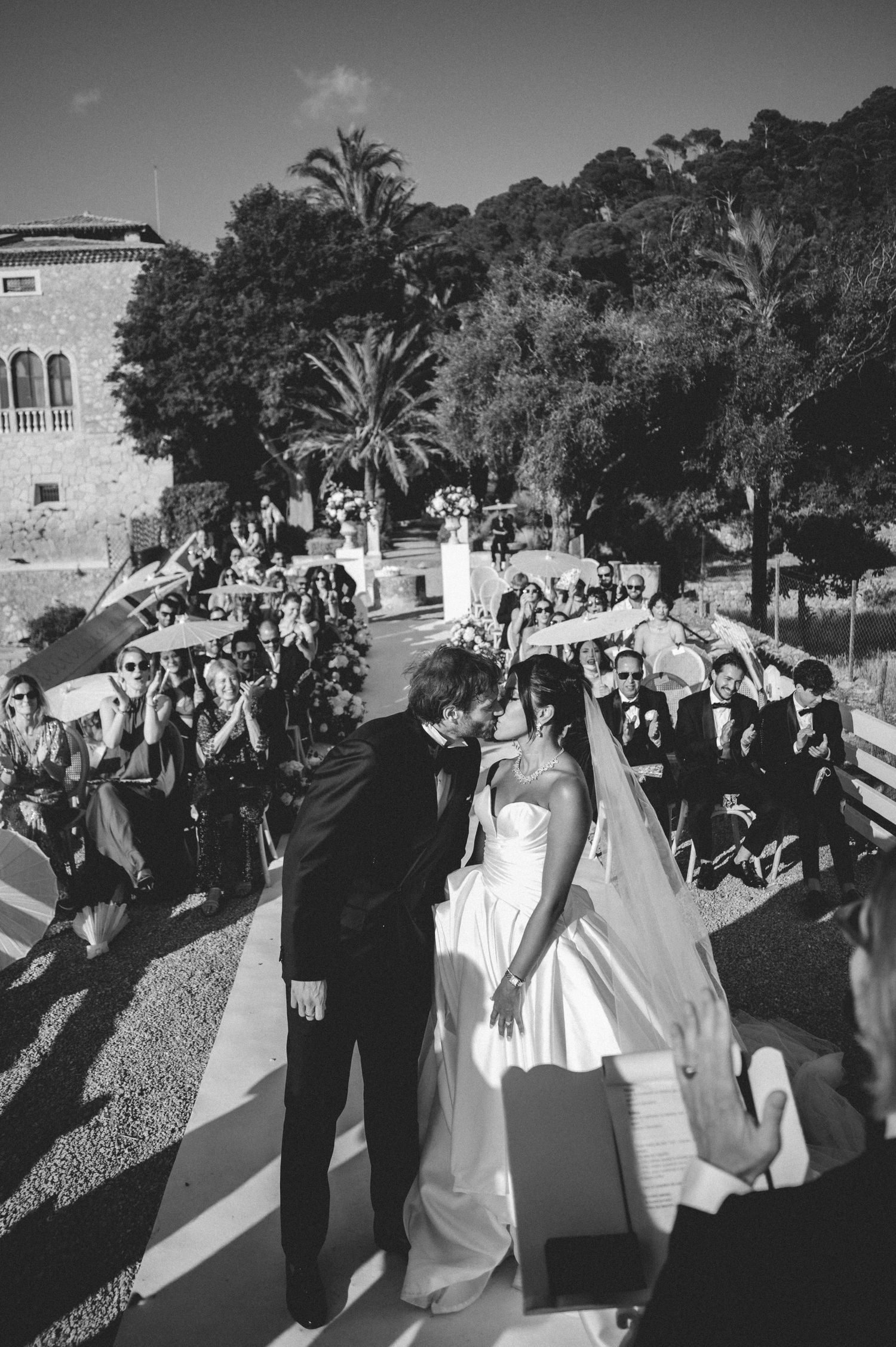 Black and white photo of groom kissing bride as guests cheer along the wedding aisle outdoors.