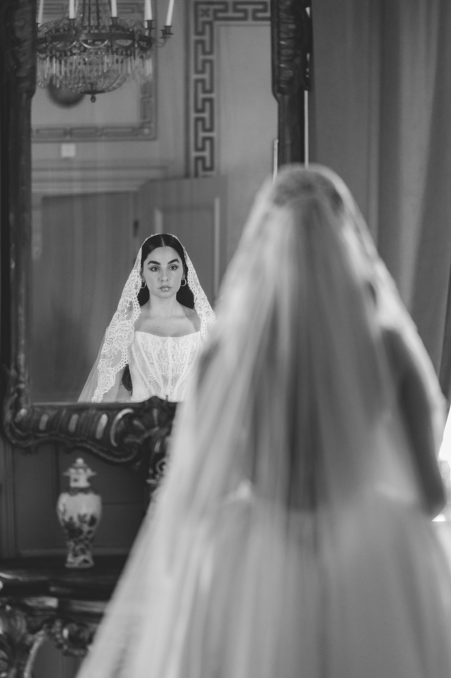 Black and white photo of bride in lace veil and wedding gown viewing her reflection in ornate mirror.