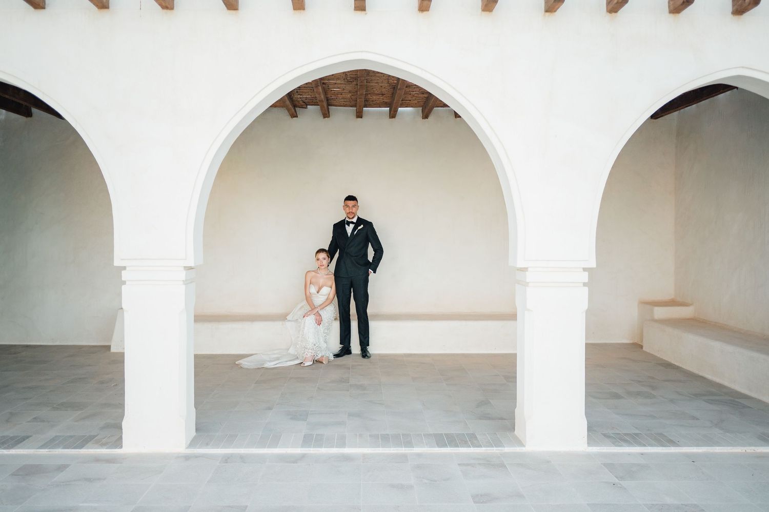 Bride and groom stand under white arched colonnade at elegant outdoor wedding venue with rustic wooden ceiling.