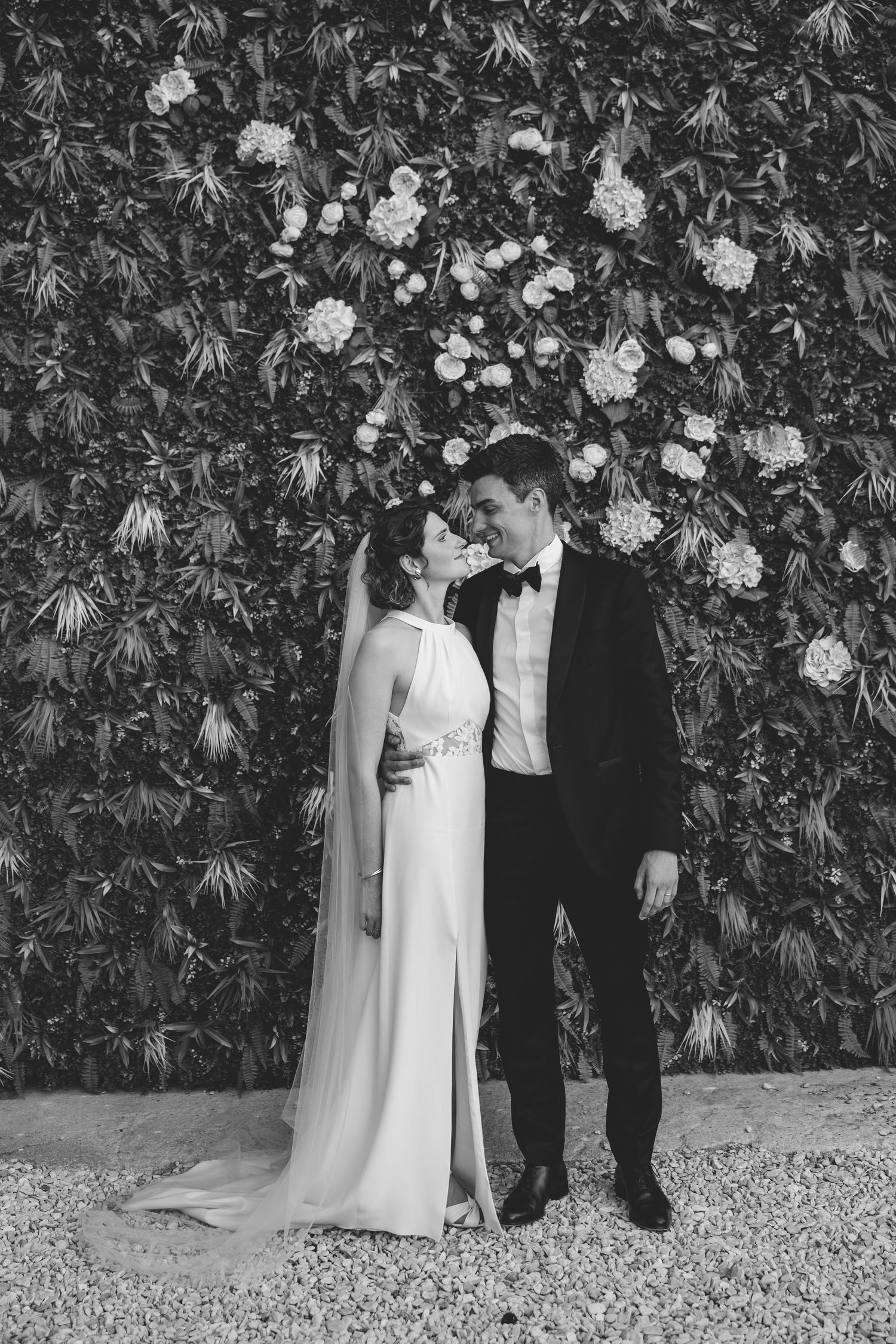 Bride and groom share a kiss before floral greenery wall in black and white photo.