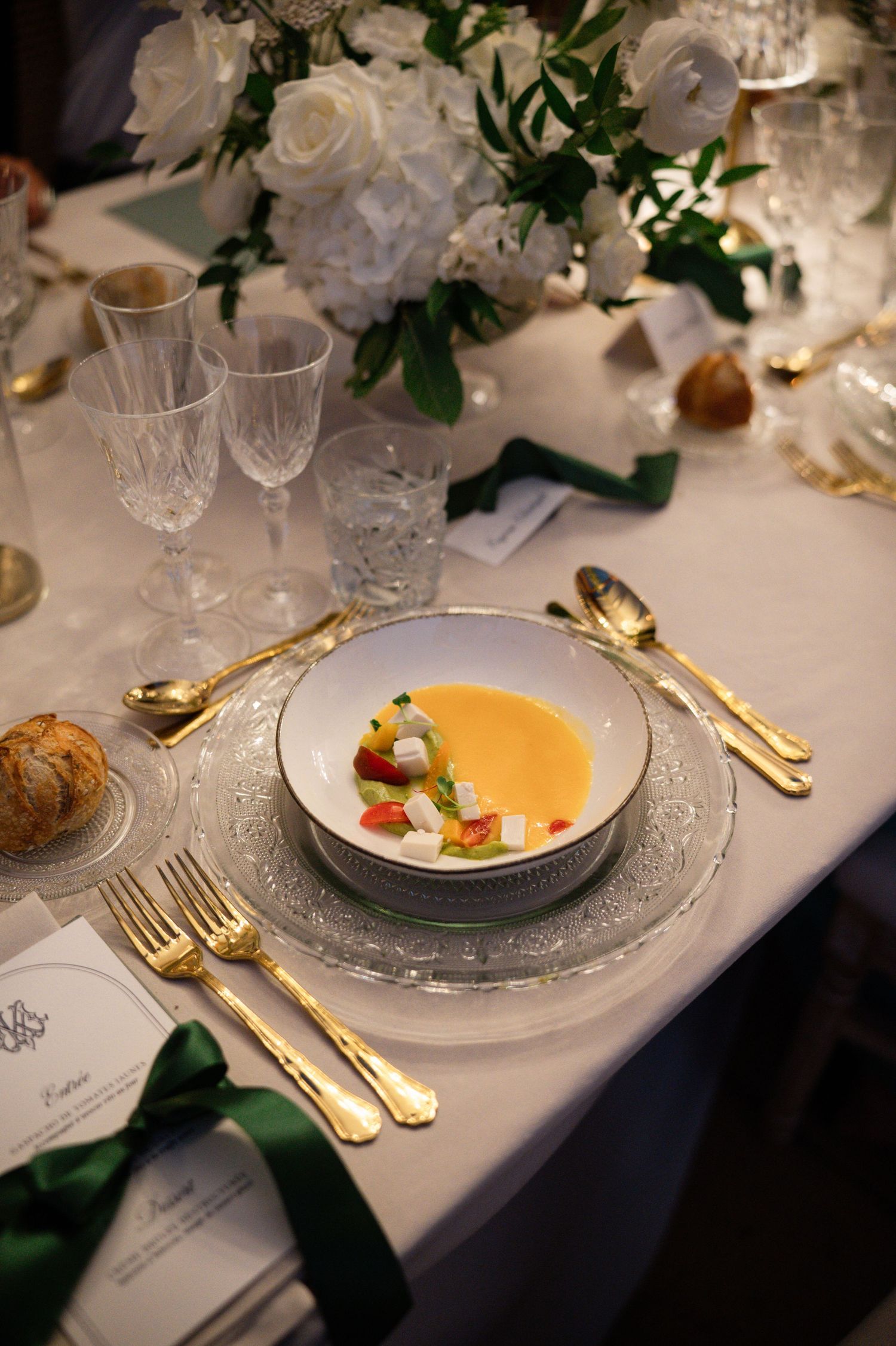 Elegant wedding table setting with gold cutlery, crystal glasses, white bowl of yellow soup, and green ribbon accents.