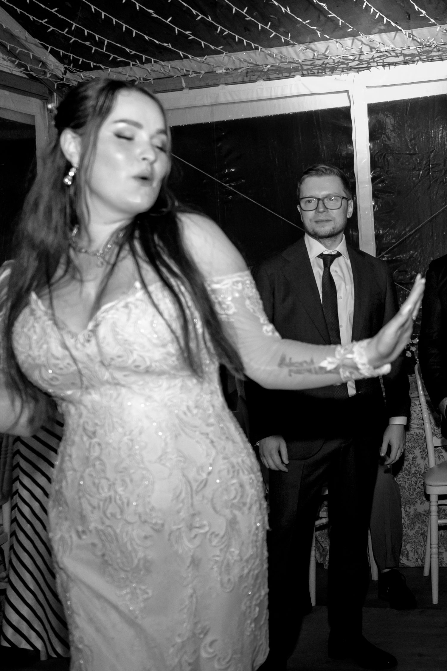 Bride in lace wedding dress dancing at reception while groom in black suit watches in black and white photo.