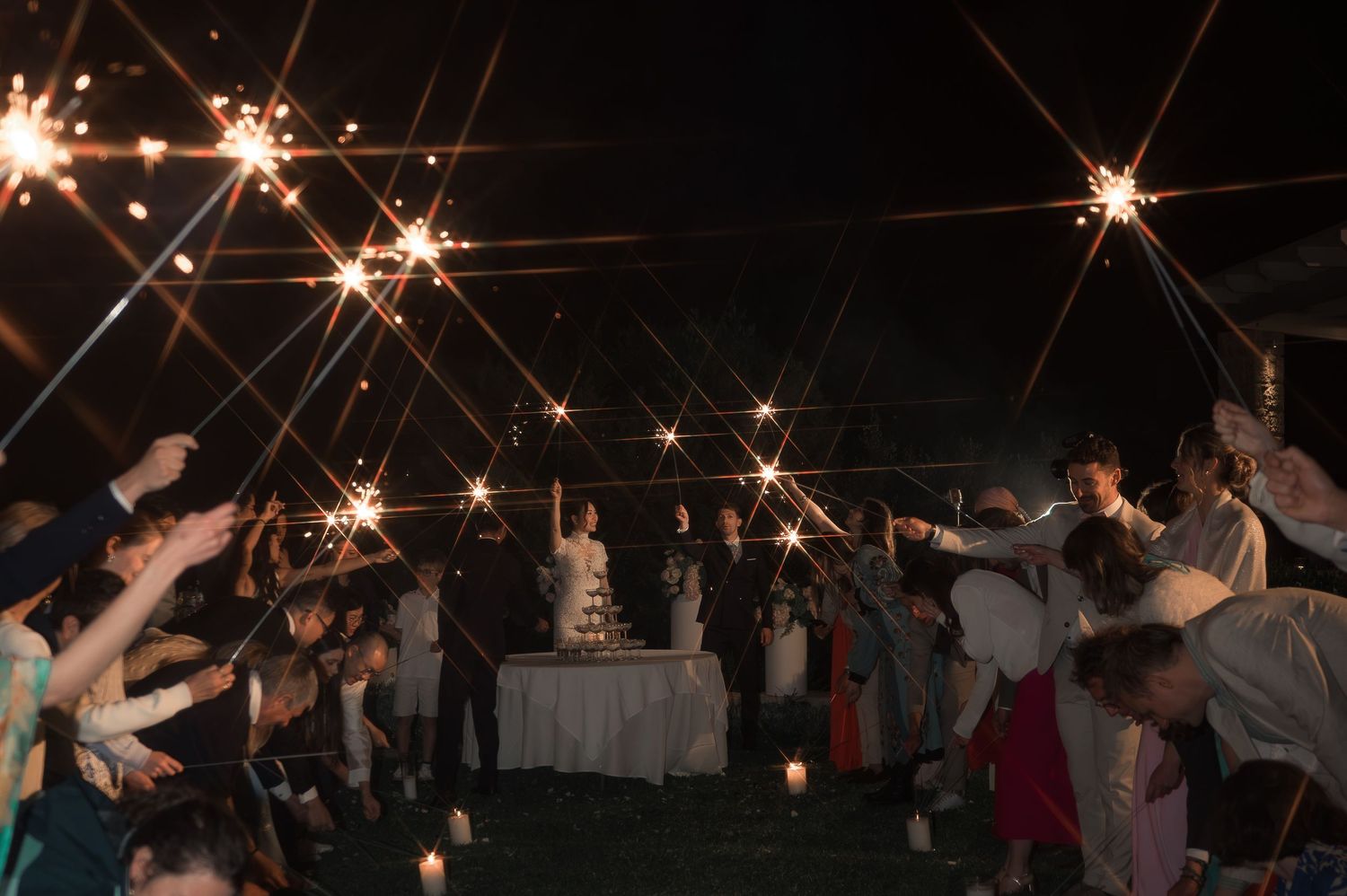 Wedding guests holding sparklers around cake table during outdoor evening wedding celebration.
