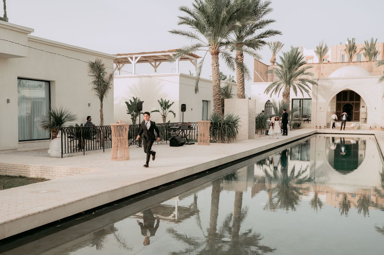 Groom in black suit running along reflective pool at luxury Moroccan wedding venue.