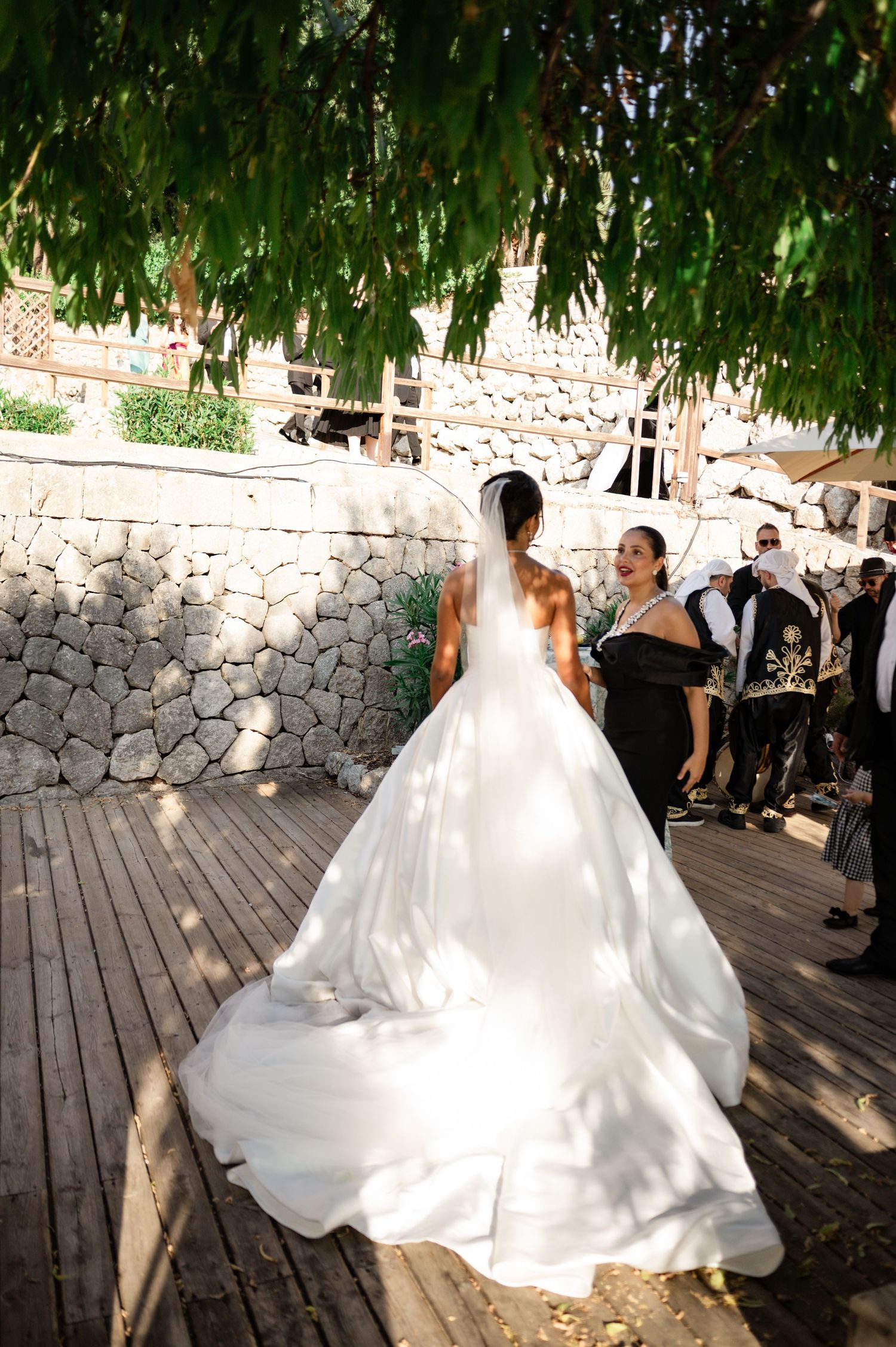 Bride in white ball gown with cathedral veil walks on wooden deck under lush green tree.