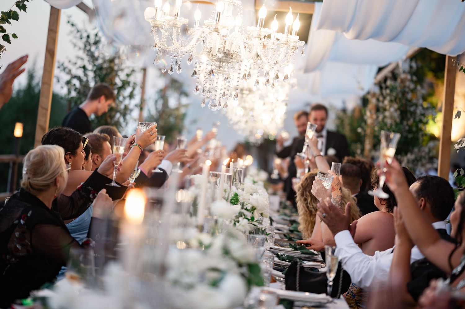 Wedding guests raise champagne glasses in toast under crystal chandeliers at elegant reception dinner.