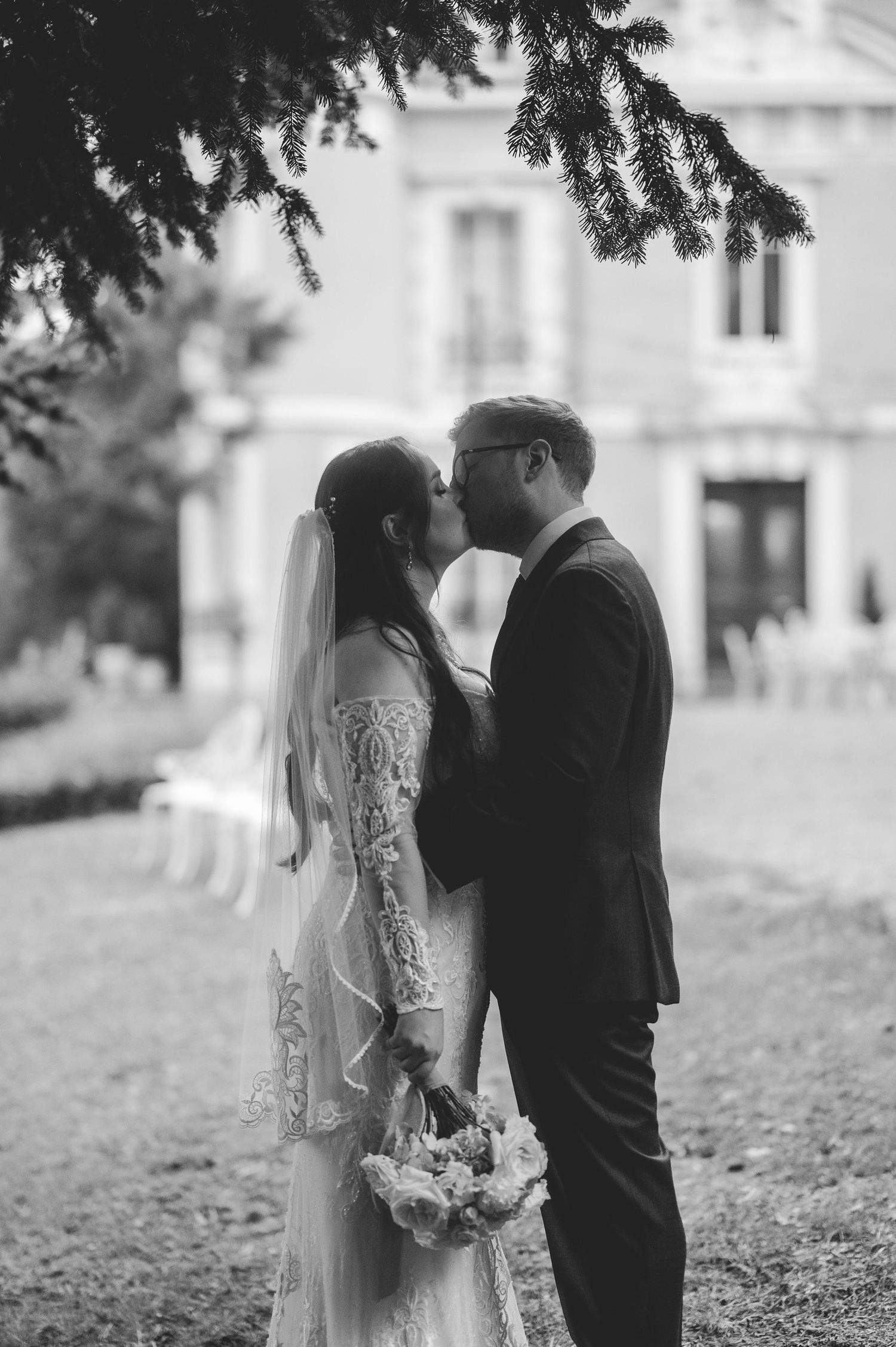 Black and white photo of bride and groom kissing in chateau garden, bride holding bouquet.