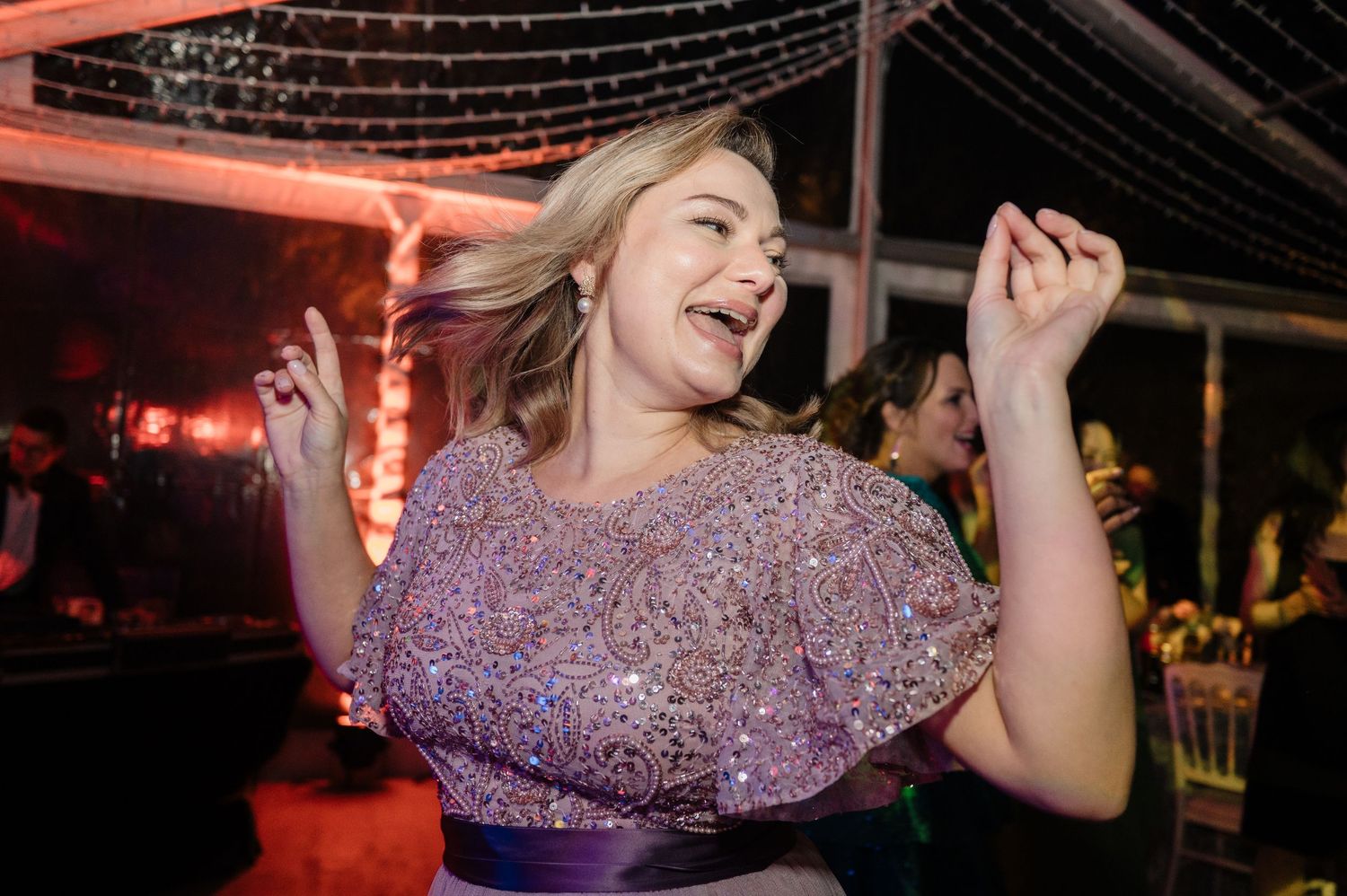 Guest in sparkly lavender dress dances joyfully at wedding reception under festive lights.