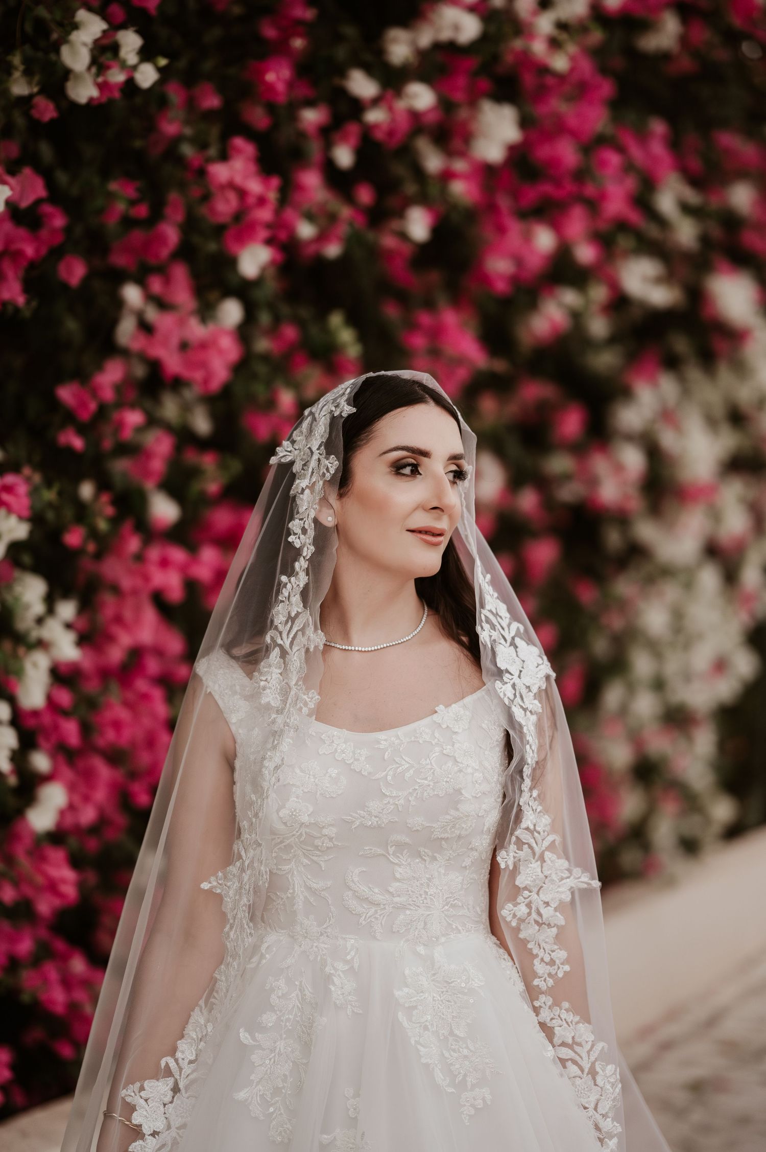 Beautiful bride in lace wedding gown and veil poses in front of vibrant pink and white bougainvillea.