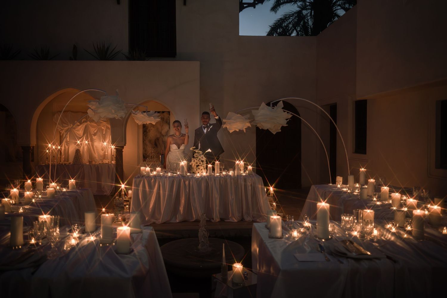 Romantic candlelit wedding reception with glowing tables and couple standing at sweetheart table with feather decorations.