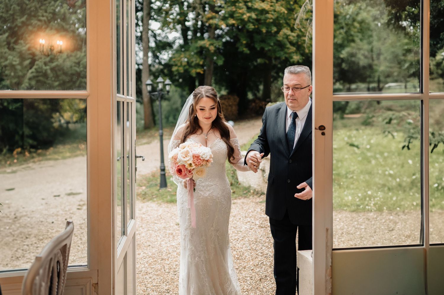 Bride in lace gown holding bouquet enters ceremony venue through French doors escorted by her father.