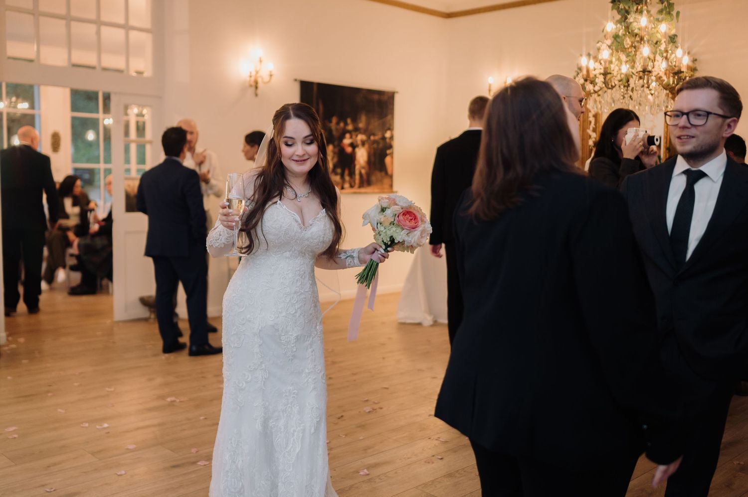 Bride in lace gown holding floral bouquet conversing with guest at elegant reception.
