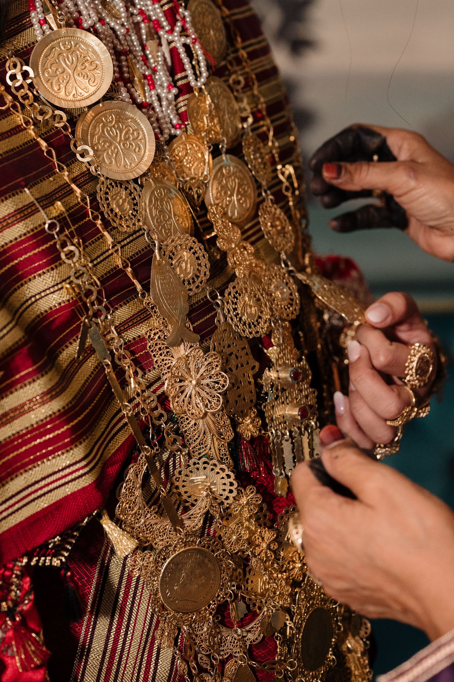 Hands adjusting ornate gold coin jewelry on a traditional striped ceremonial garment.
