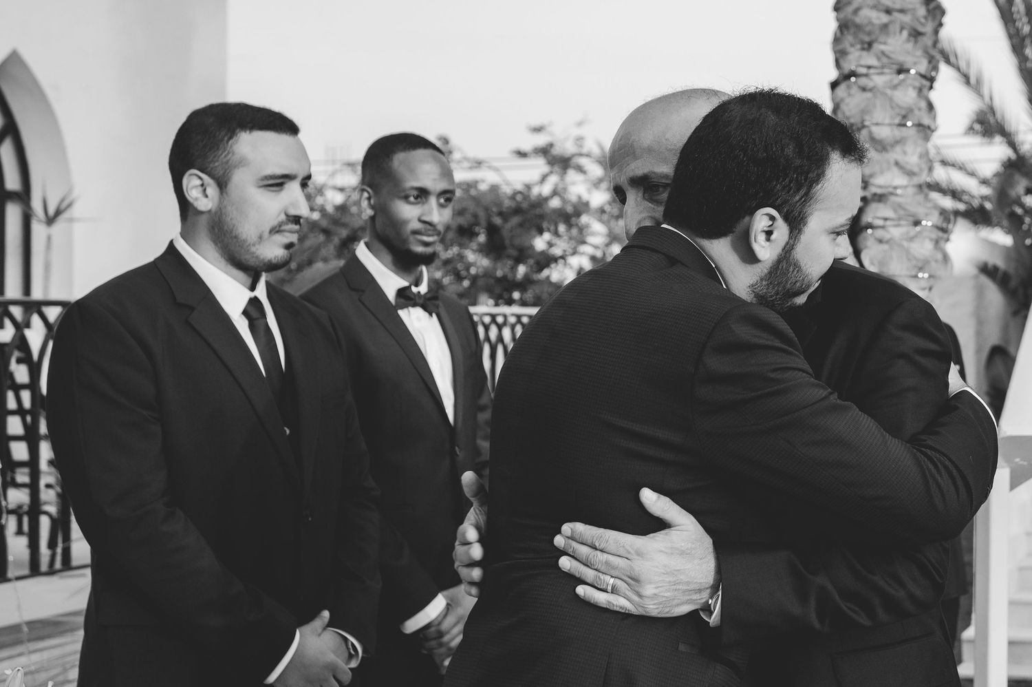 Groomsmen in black suits sharing an emotional embrace while two others look on at an outdoor wedding venue.