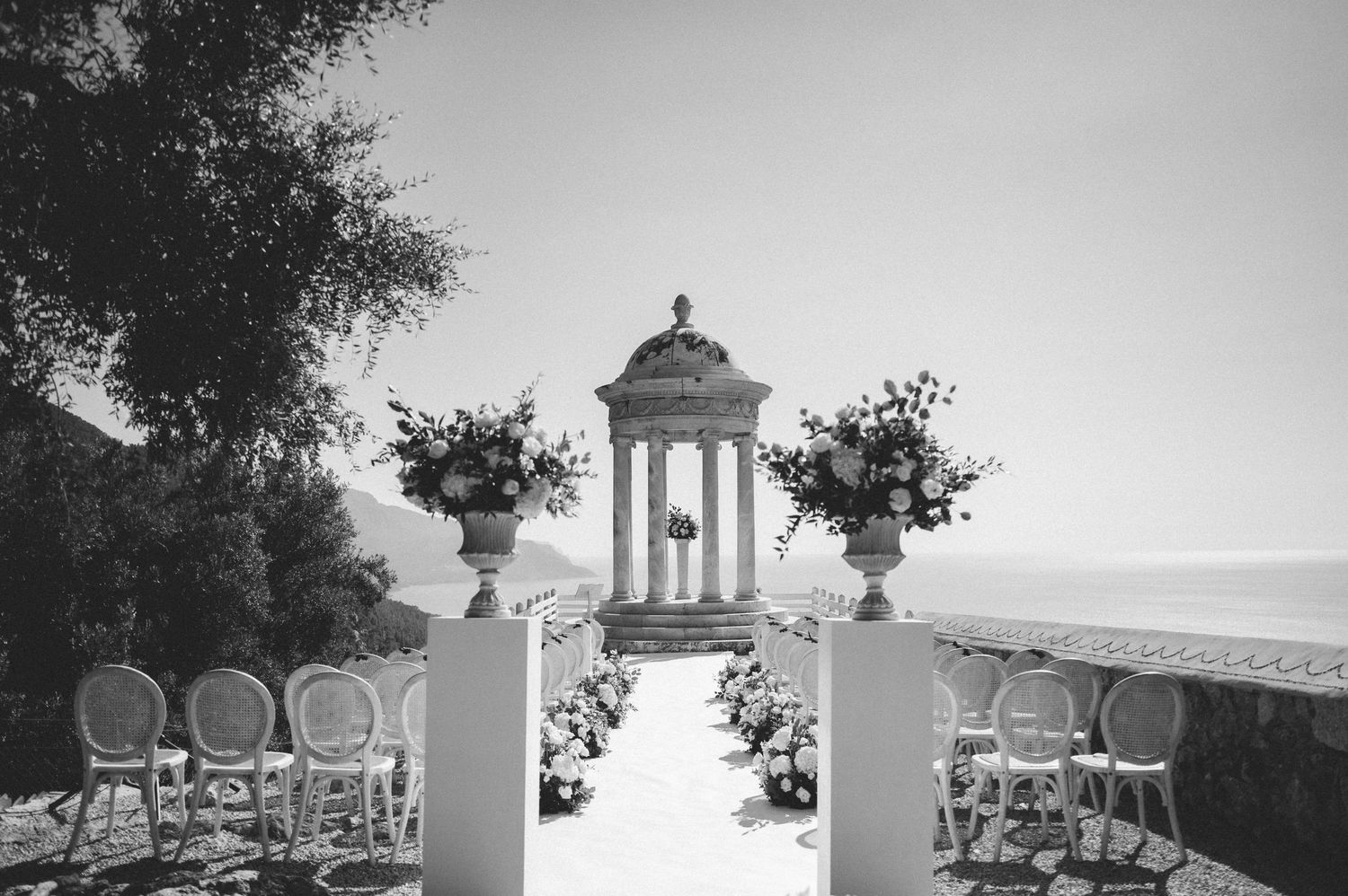 Black and white photo of an outdoor wedding ceremony setup with a classical gazebo and floral arrangements by the sea.