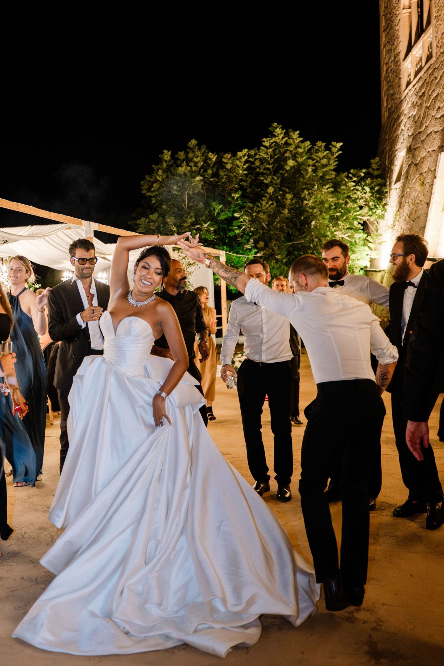 Radiant bride in voluminous white satin gown dancing joyfully with guests at outdoor nighttime wedding celebration.
