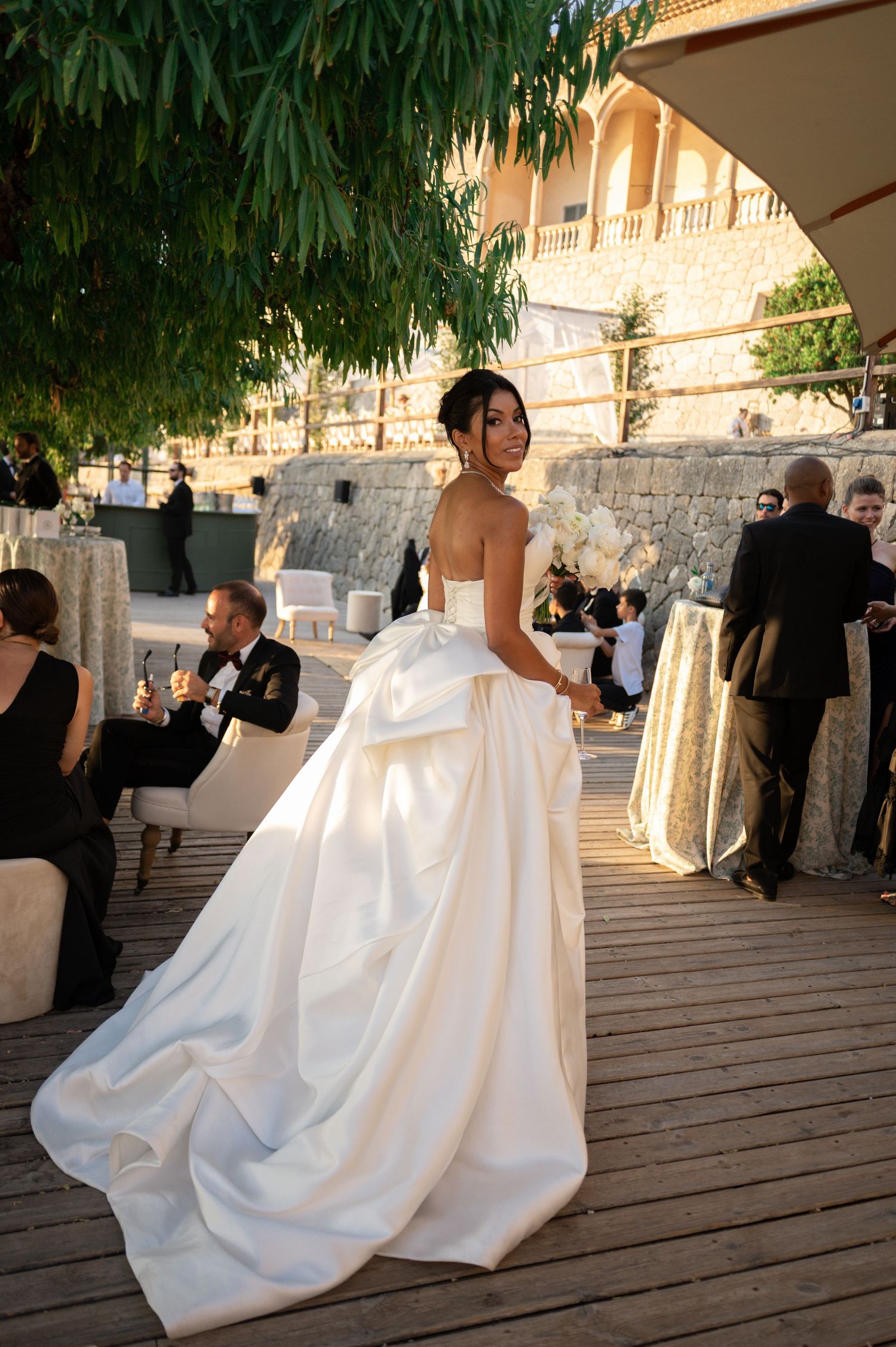 Elegant bride in strapless white satin ball gown with dramatic bow back at outdoor Mediterranean wedding venue.