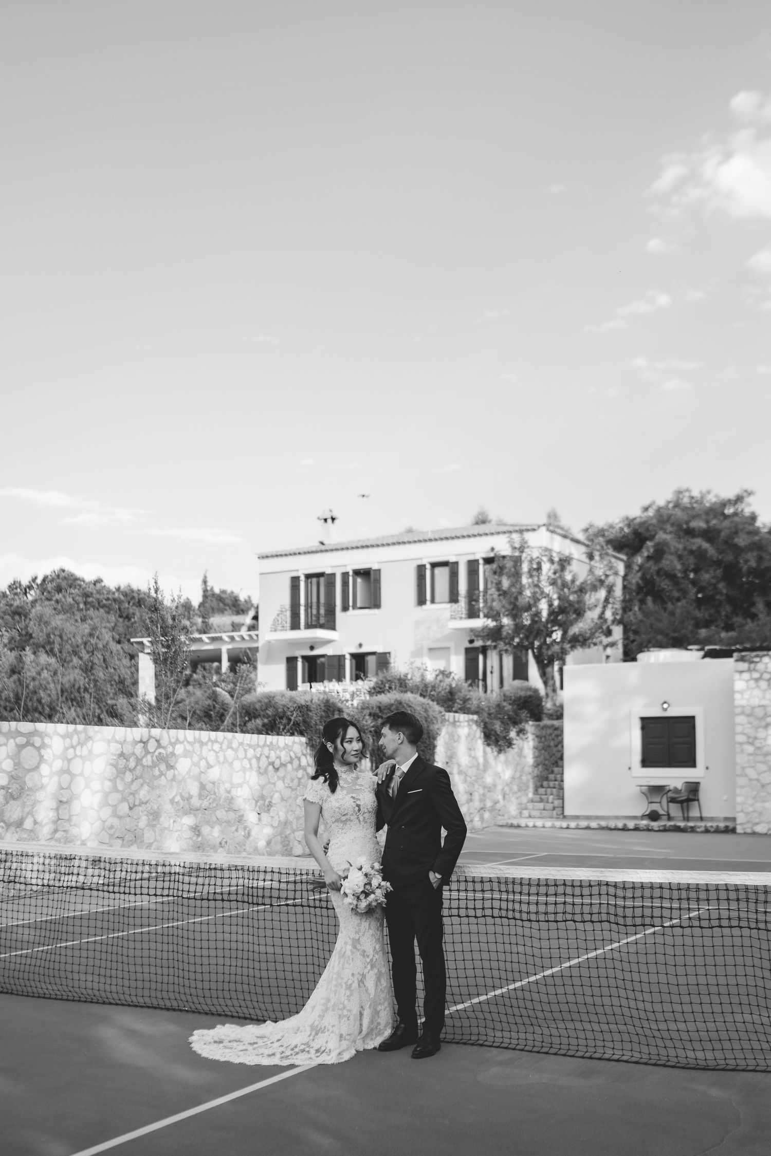 Bride and groom share a romantic moment together at a tennis court net with a villa in the background.