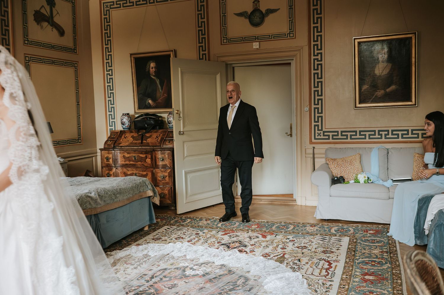 Man in dark suit entering elegant room where bride in white gown waits near ornate portrait-lined walls.