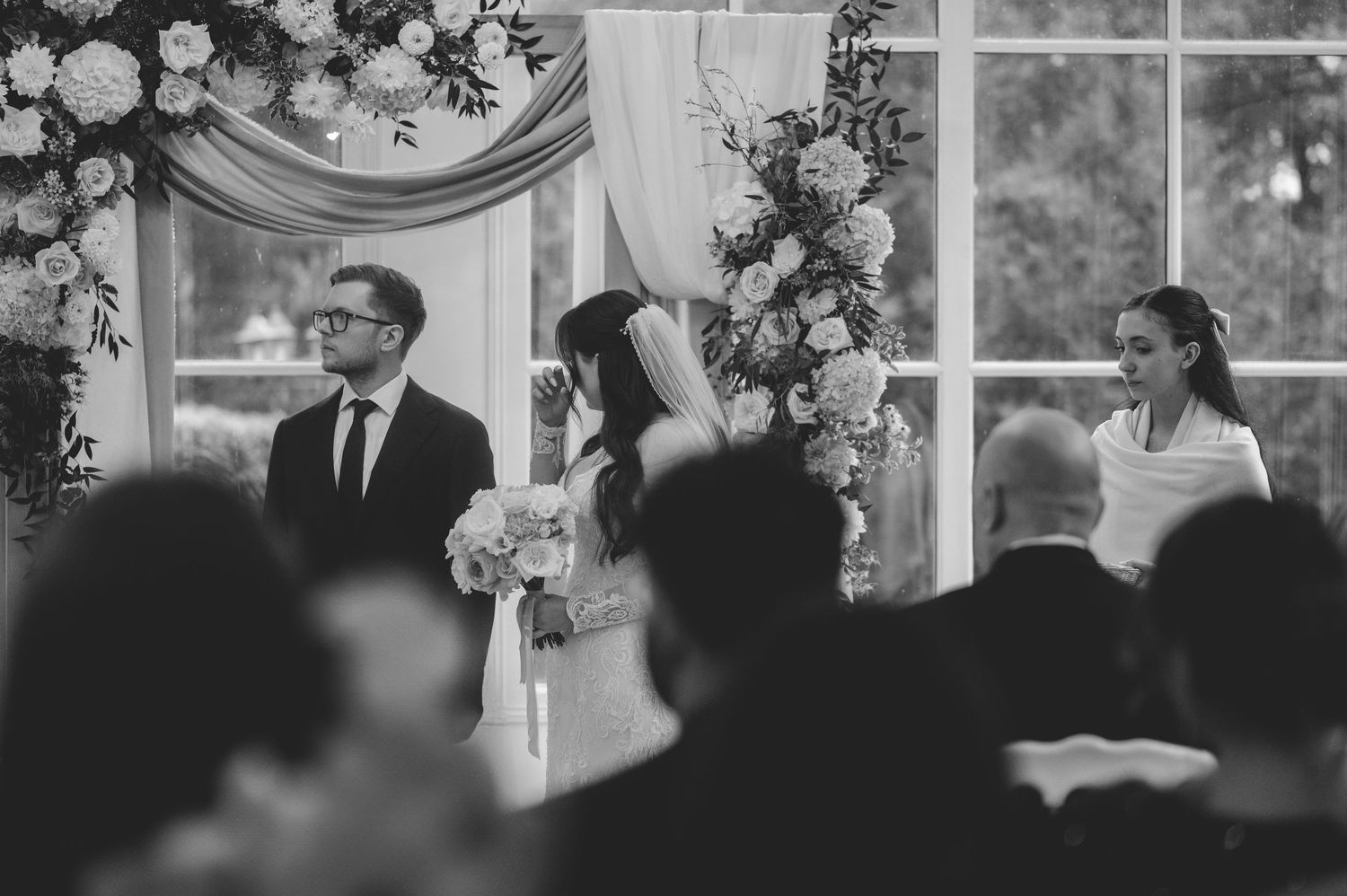 Bride and groom stand beneath floral arch during wedding ceremony in black and white photo.