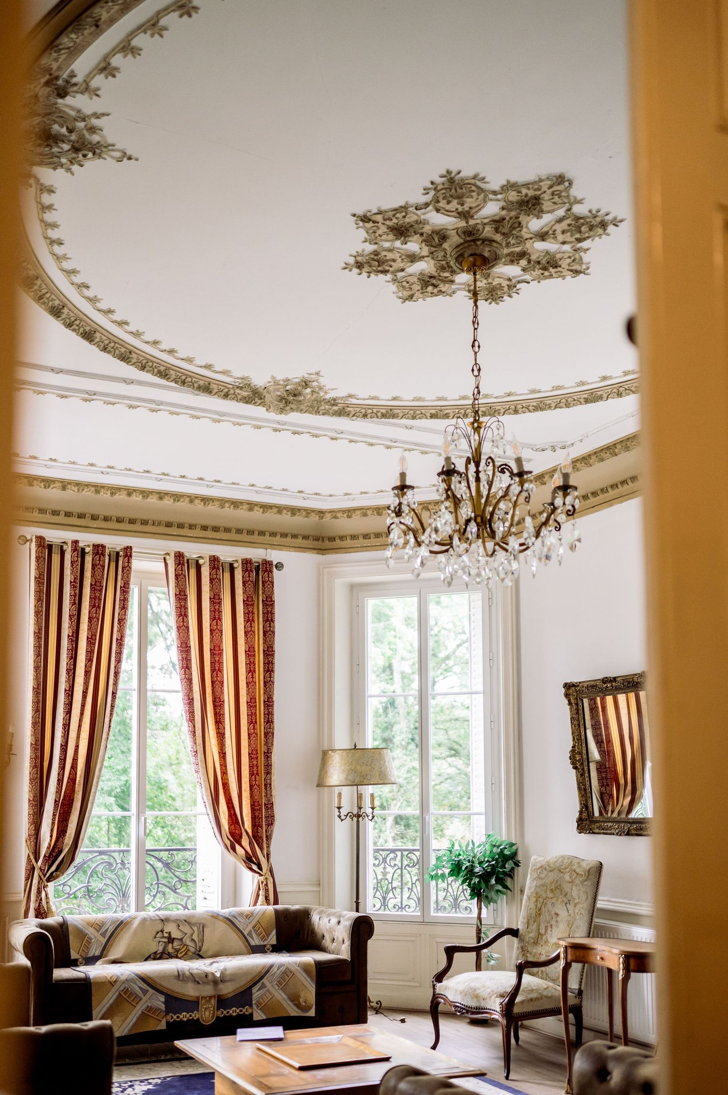 Ornate French chateau salon with gilded ceiling medallion, crystal chandelier, and rich red damask curtains.