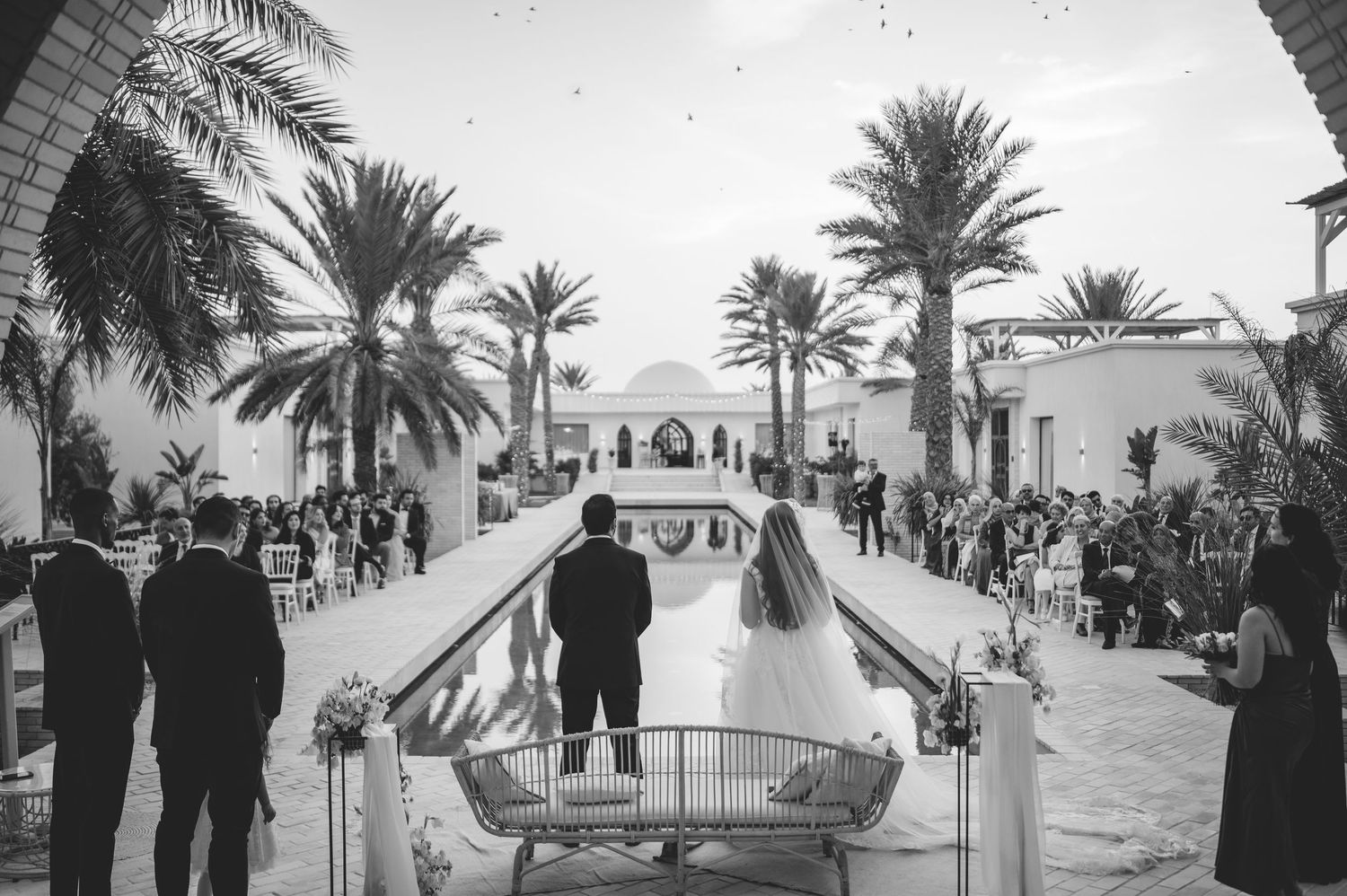Black and white aerial view of bride and groom at outdoor wedding ceremony with reflecting pool and palm trees.