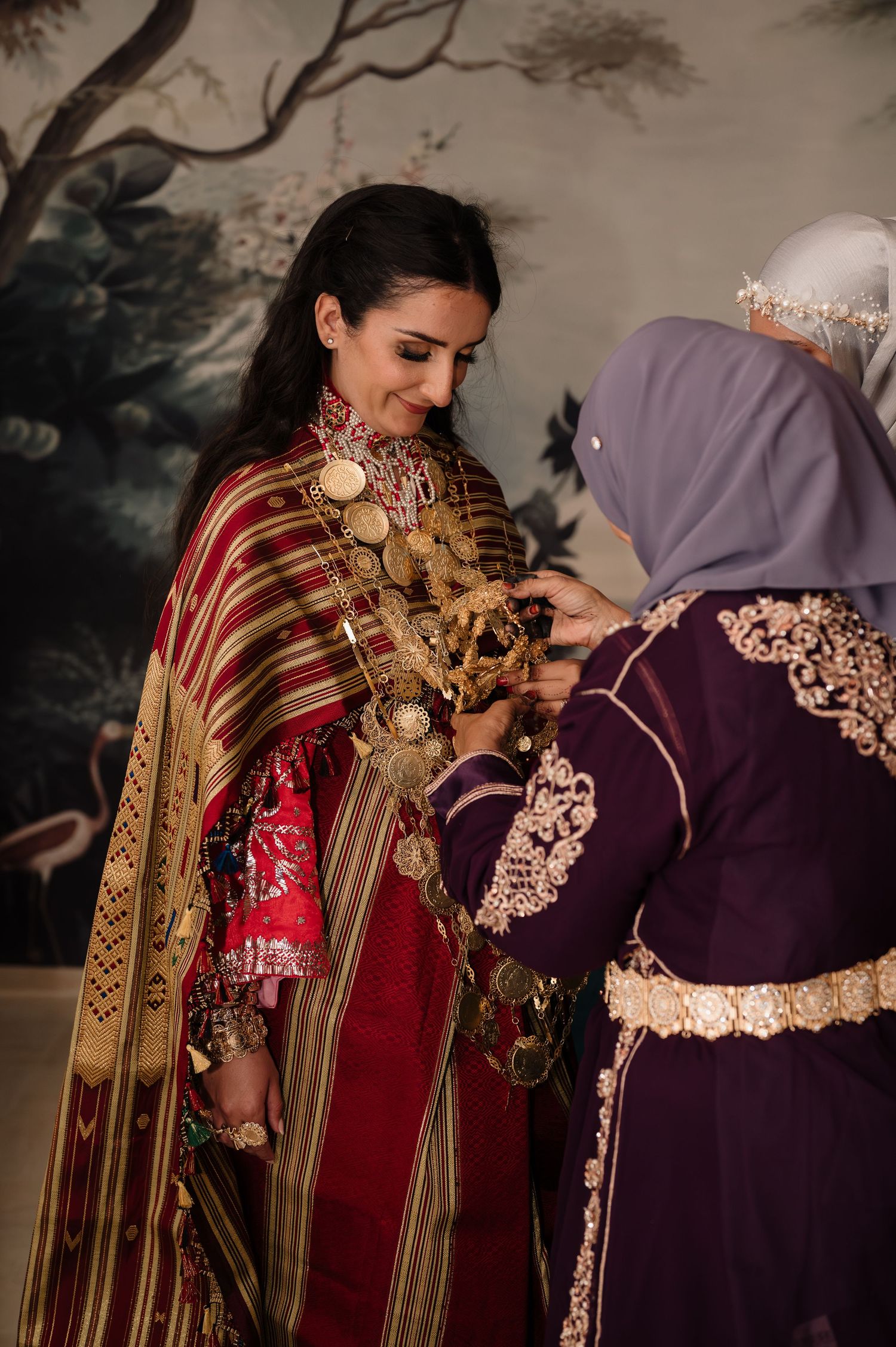 Woman being dressed in traditional red and gold bridal attire with coin jewelry.